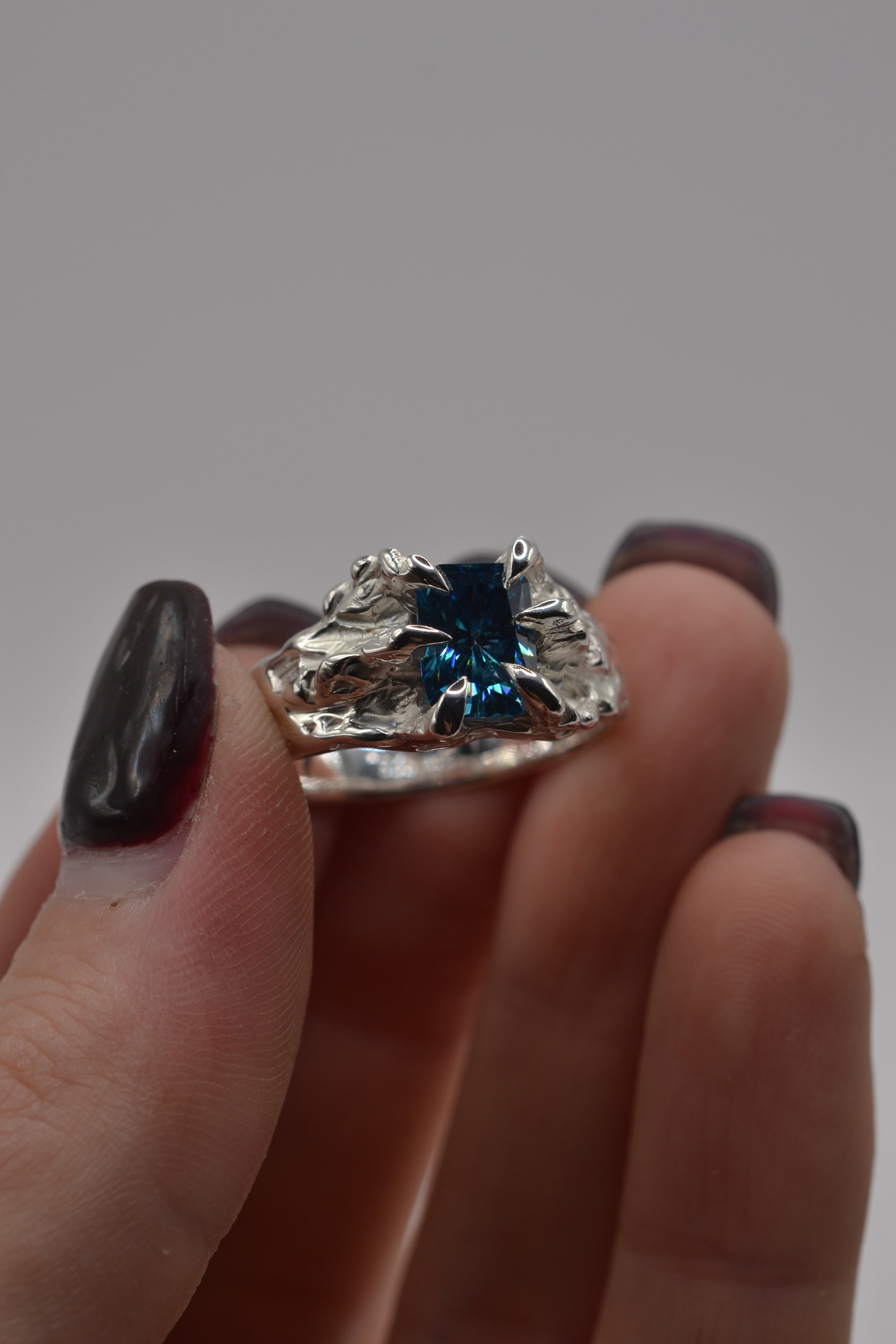Detailed view of a lost wax cast silver ring with a blue radiant cut moissanite, showcasing organic textures and claw setting.