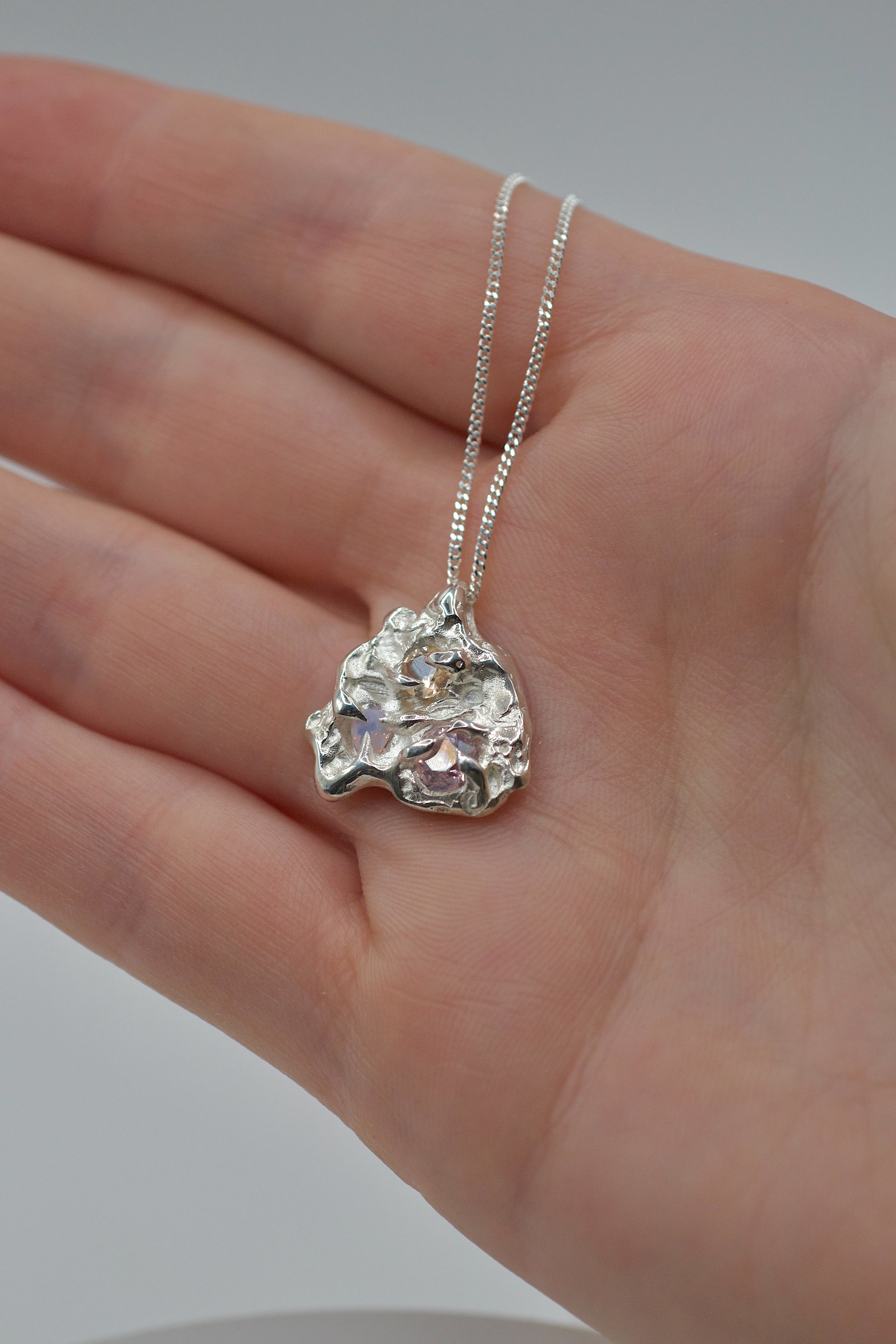 Hand holding a molten 925 sterling silver pendant with two cast-in-place lab-grown gemstones, handmade using the lost wax technique.