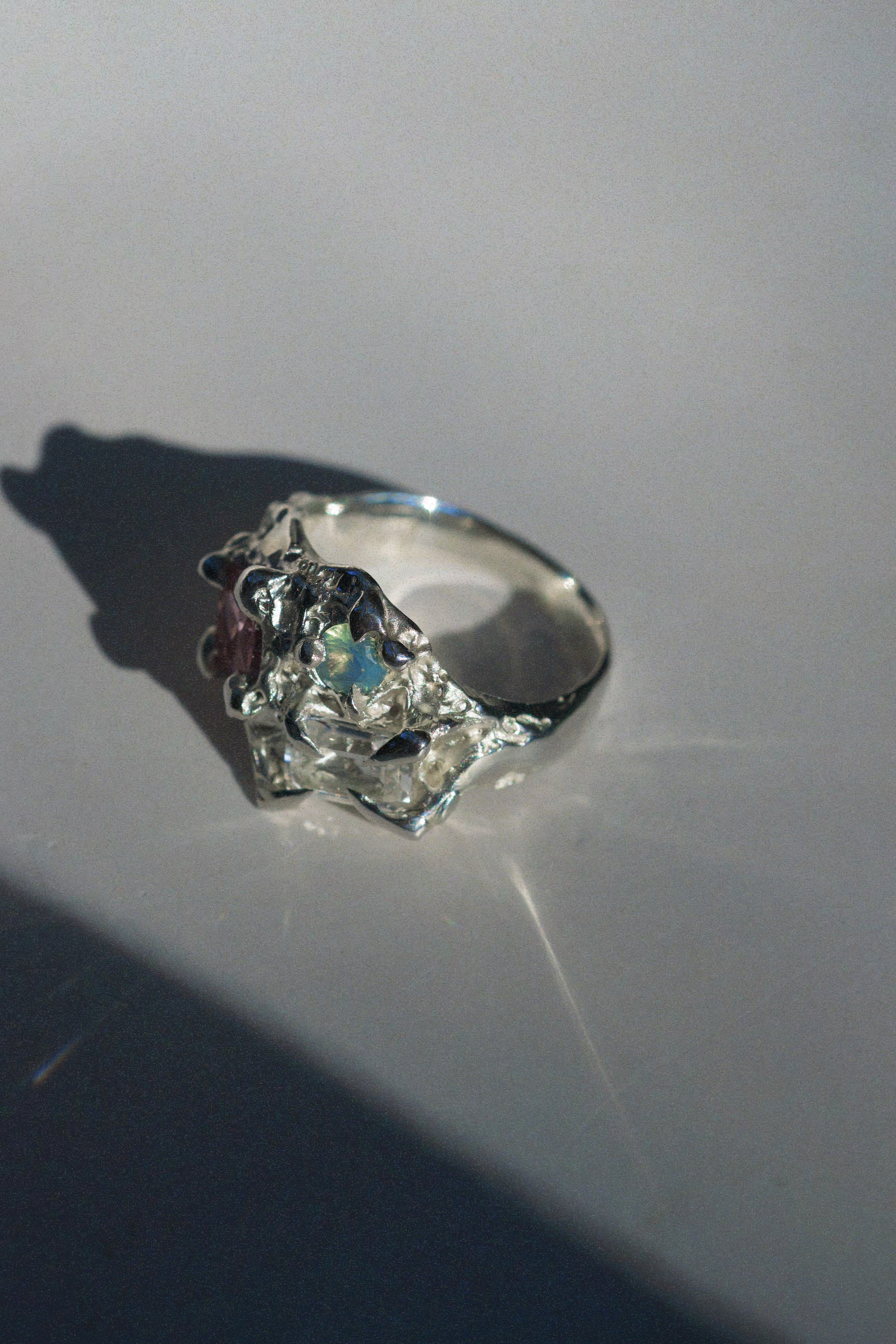 Side angle of the Wild Flower ring revealing raw, uneven silver textures and elevated prongs encasing an opal, cubic zirconia, and sapphire in asymmetrical alignment. The design resembles blooming petals and roots in metal form.