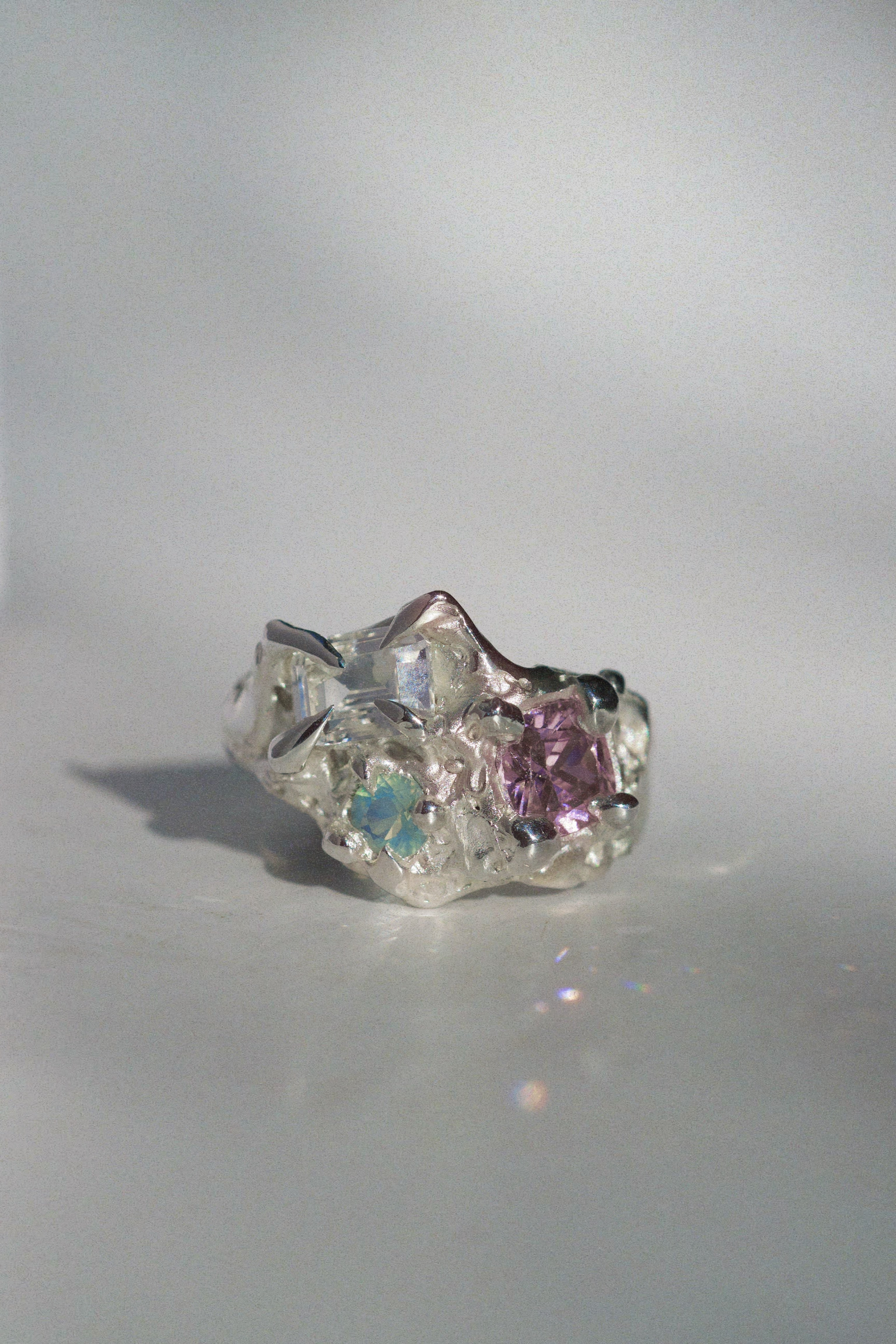 Side view of a handmade fantasy-style silver ring with a cluster gemstone setting, showcasing an emerald-cut clear white sapphire, a cushion-cut pink cubic zirconia, and a round lab-grown aqua opal. The ring features flowing, molten textures created using lost wax casting.