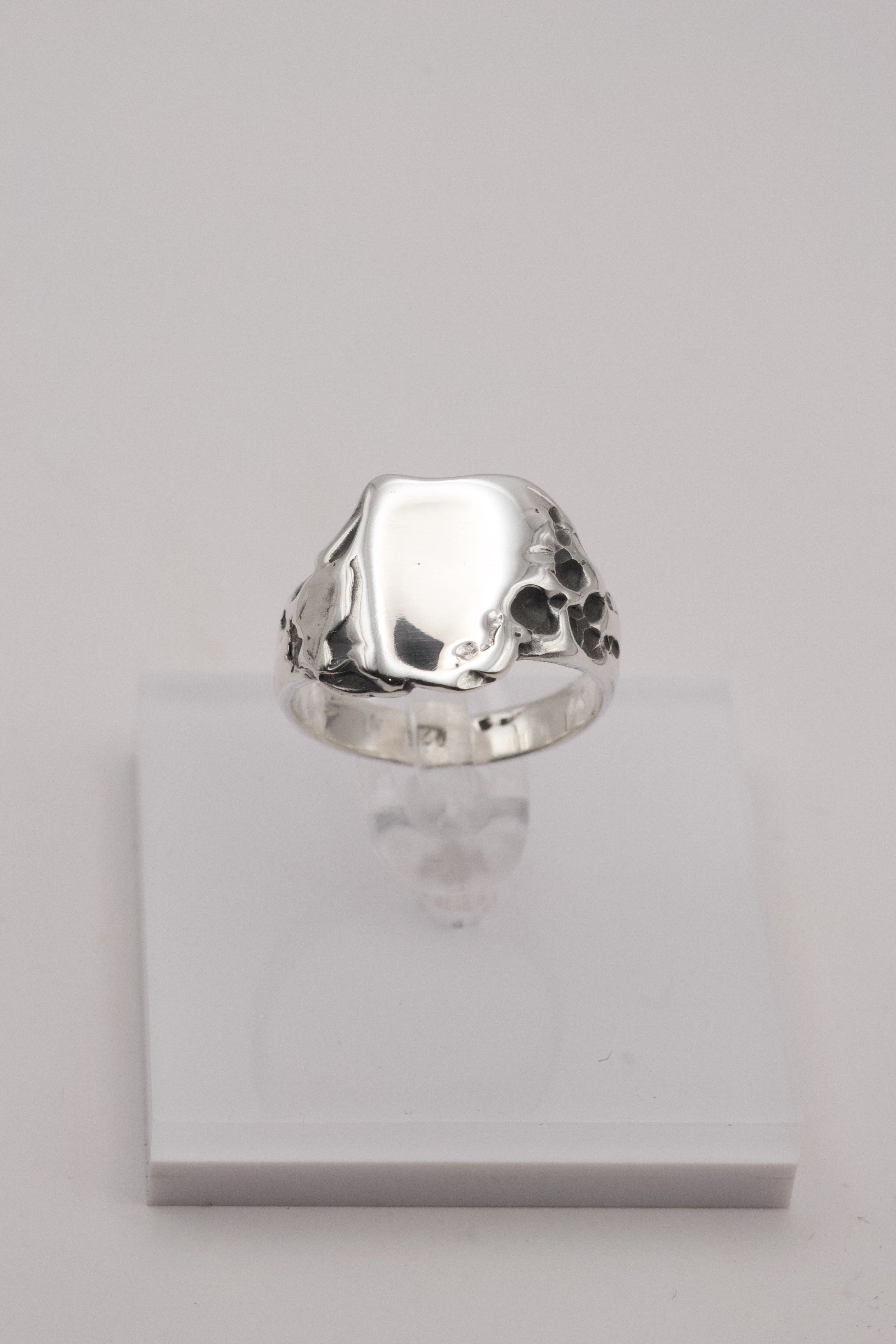 A polished silver signet ring shown upright on a white stand, highlighting its organic lost-wax-formed texture and molten carved edges.