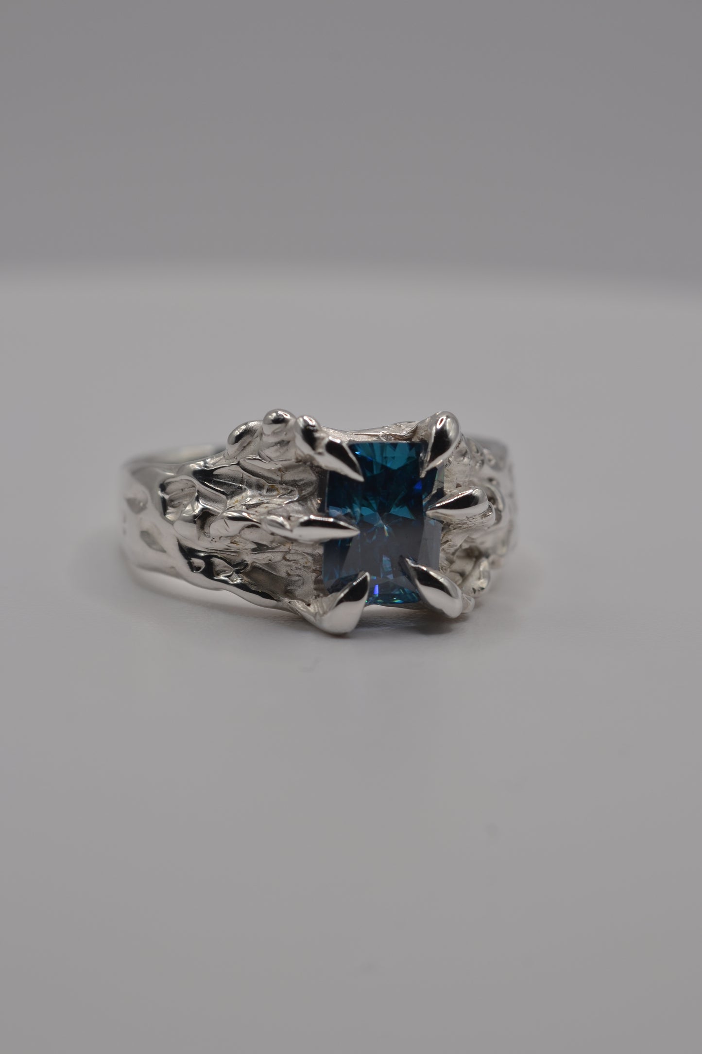 Macro detail of a blue radiant cut moissanite secured in an organic claw setting on a lost wax cast silver ring.