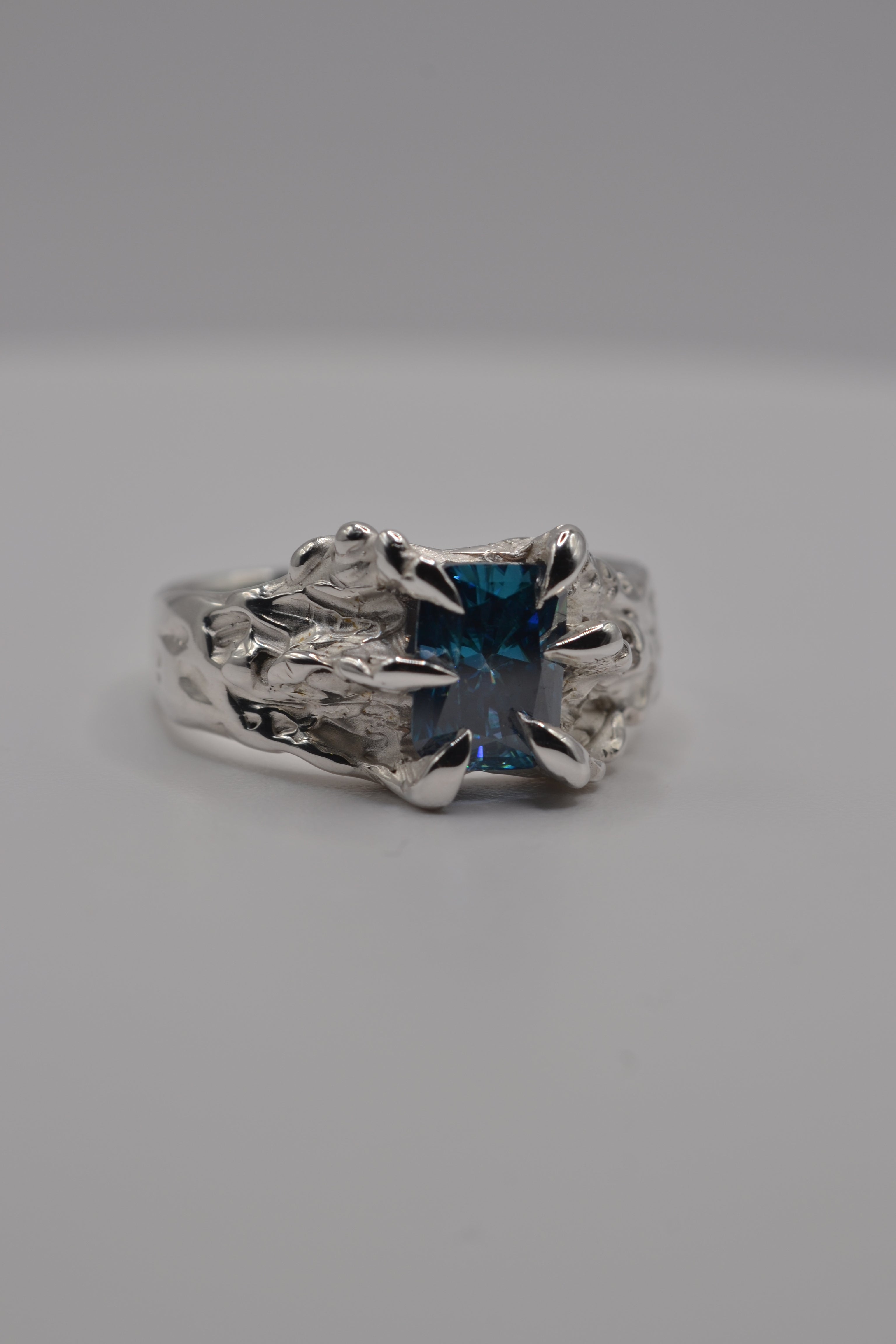 Macro detail of a blue radiant cut moissanite secured in an organic claw setting on a lost wax cast silver ring.