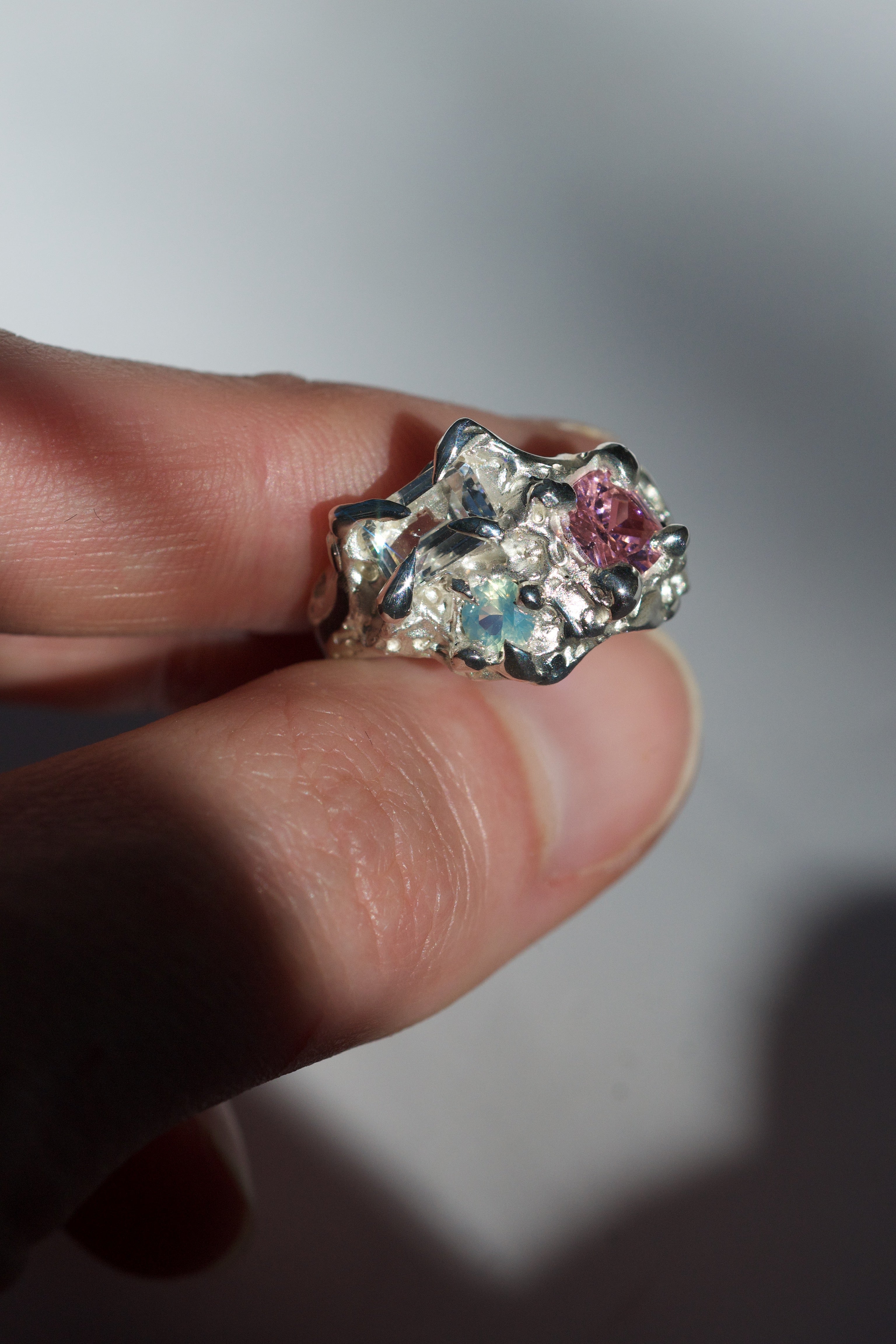 Top view of a lost wax cast silver ring showcasing three bezel-set stones: a clear emerald-cut sapphire, a soft pink cushion-cut CZ, and a glowing aqua opal. Surrounded by sculptural, flower-like metalwork with an ethereal, fantasy jewellery aesthetic.