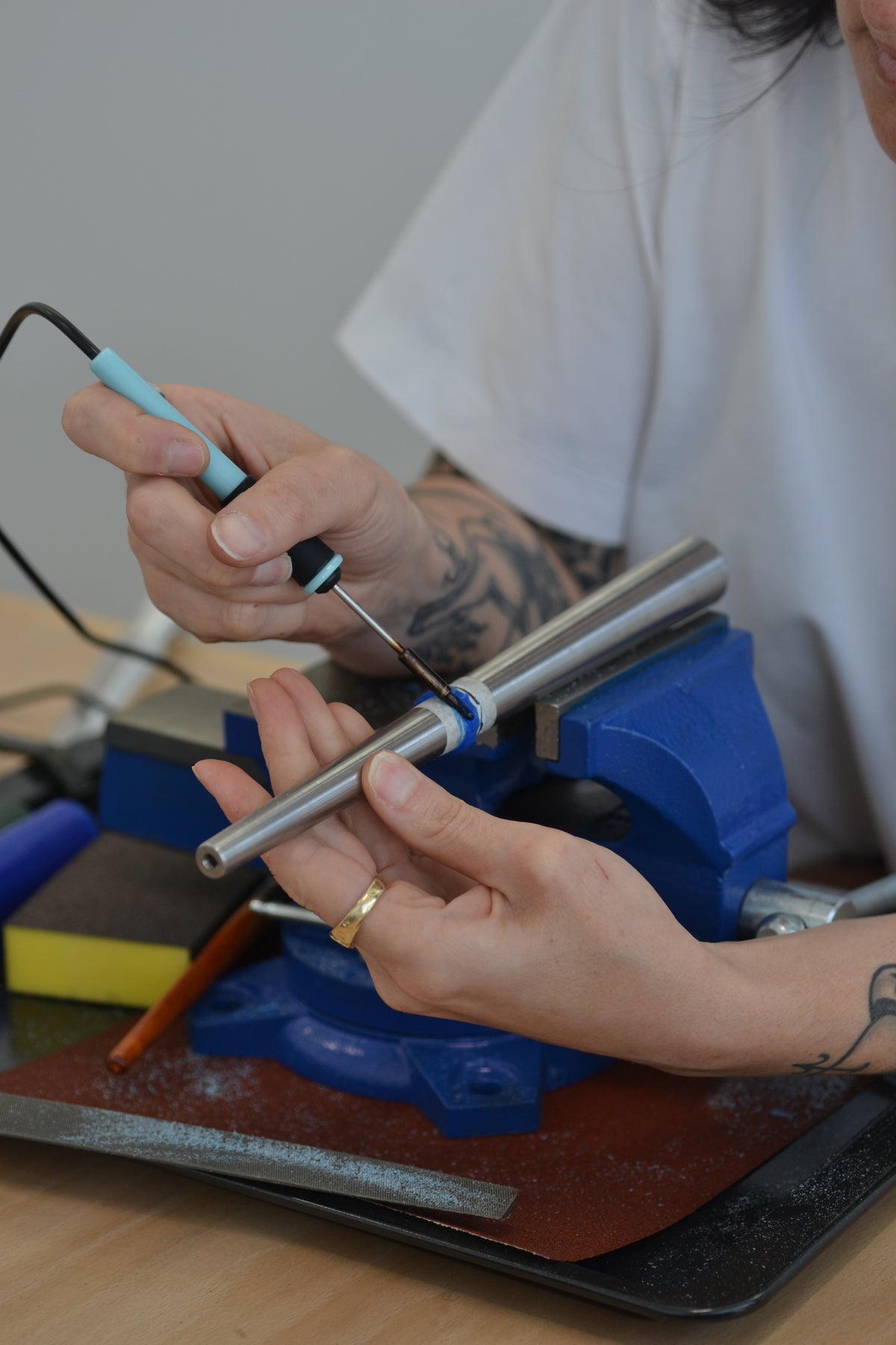 A close-up of a Cassandra Djodan's hands using a soldering iron on a metal ring mandrel on a blue bench vice, with a wax ring being sculpted.