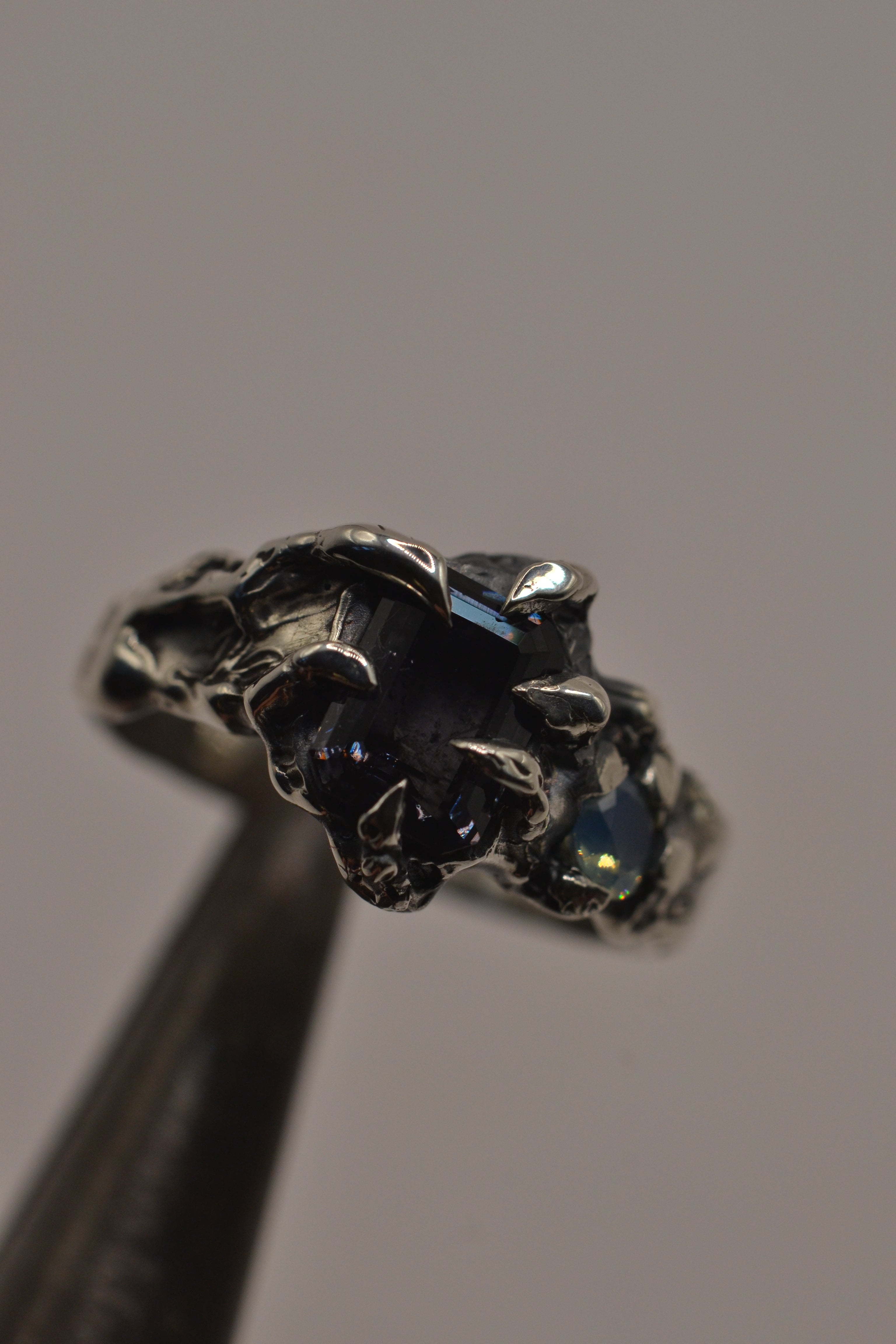 Close up of the Silhouette Ring held in tweezers showing milky aqua opal and grey spinel within darkened sterling silver claws handmade using the lost wax technique