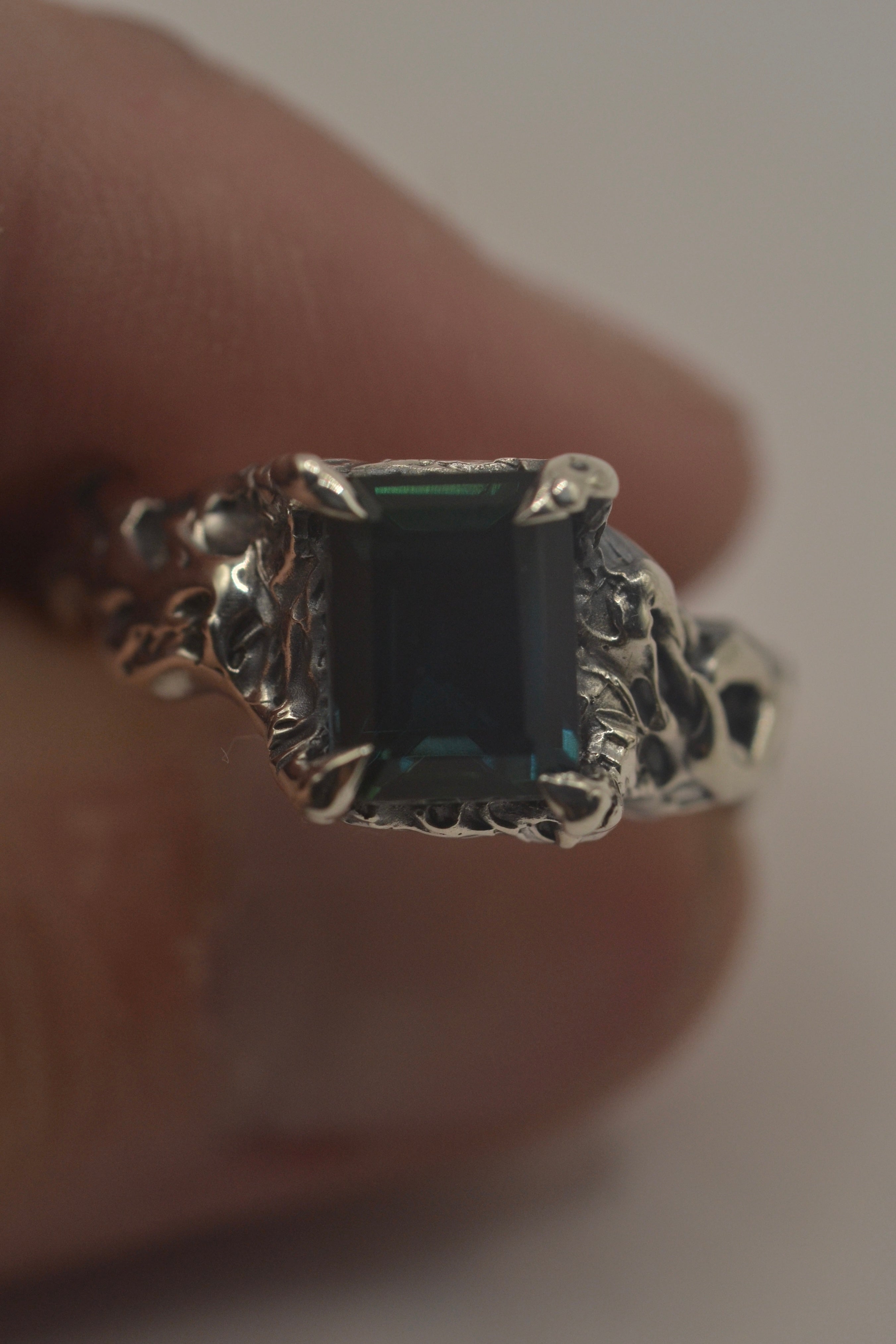 Lost wax cast darkened sterling silver Twilight Ring held im hand with teal lab-grown sapphire and heavily textured organic setting