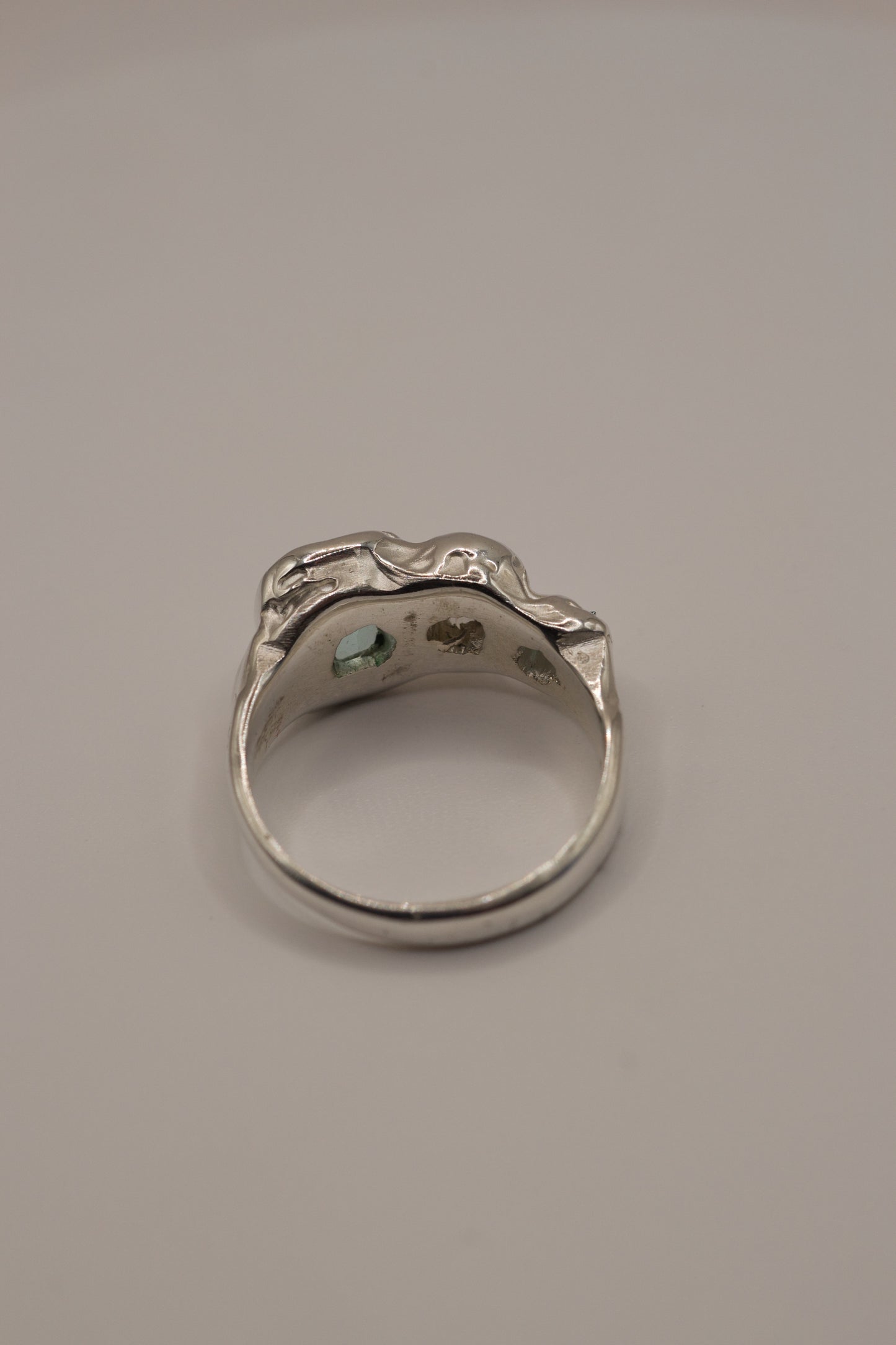 Back view of a molten textured 925 sterling silver ring with high polish surface, revealing open stone seats for cast in place gemstones: seafoam and green crystals with sparkling moissanite, all handmade through the lost wax casting process for alternative jewellery.