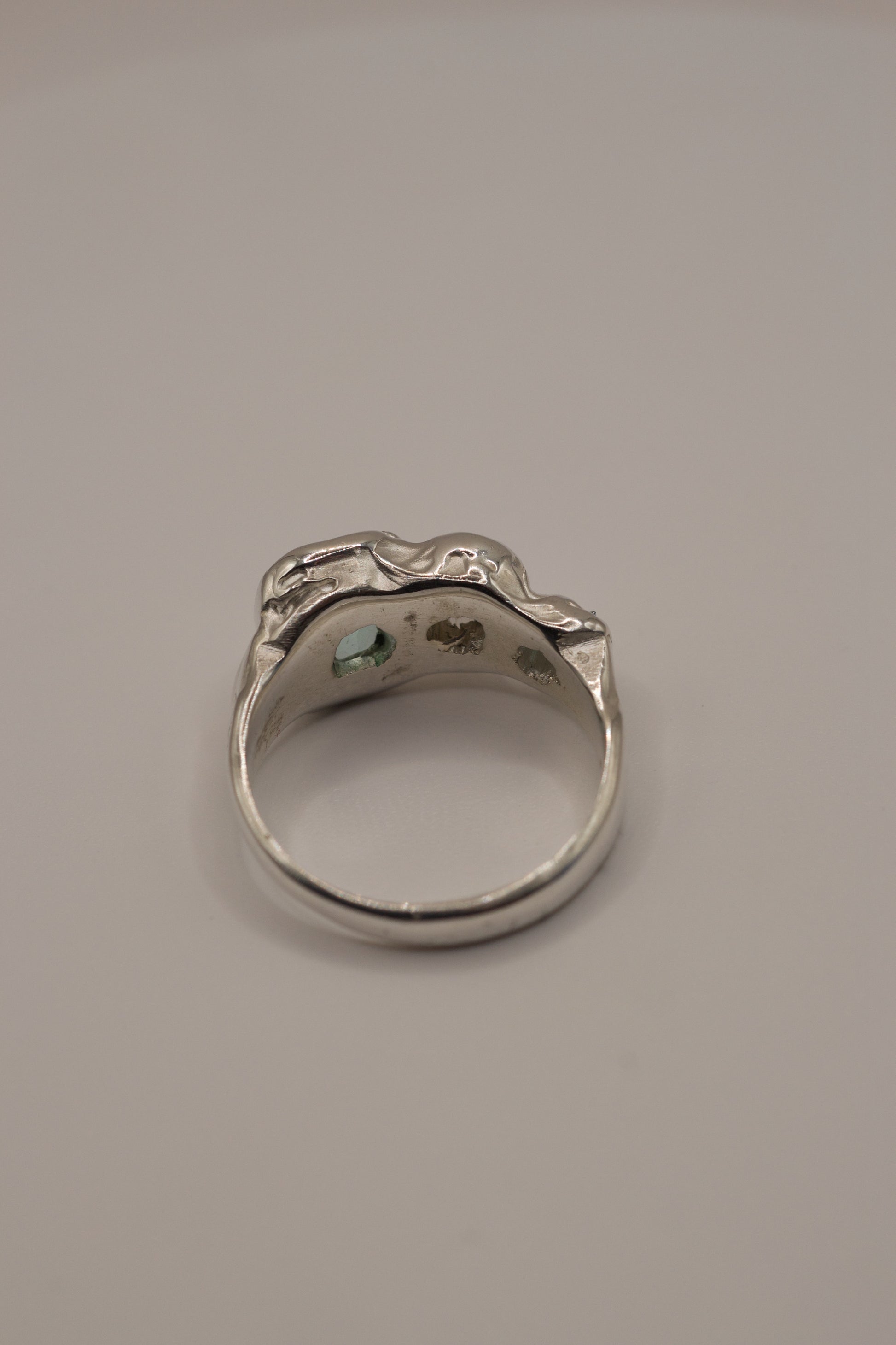 Back view of a molten textured 925 sterling silver ring with high polish surface, revealing open stone seats for cast in place gemstones: seafoam and green crystals with sparkling moissanite, all handmade through the lost wax casting process for alternative jewellery.