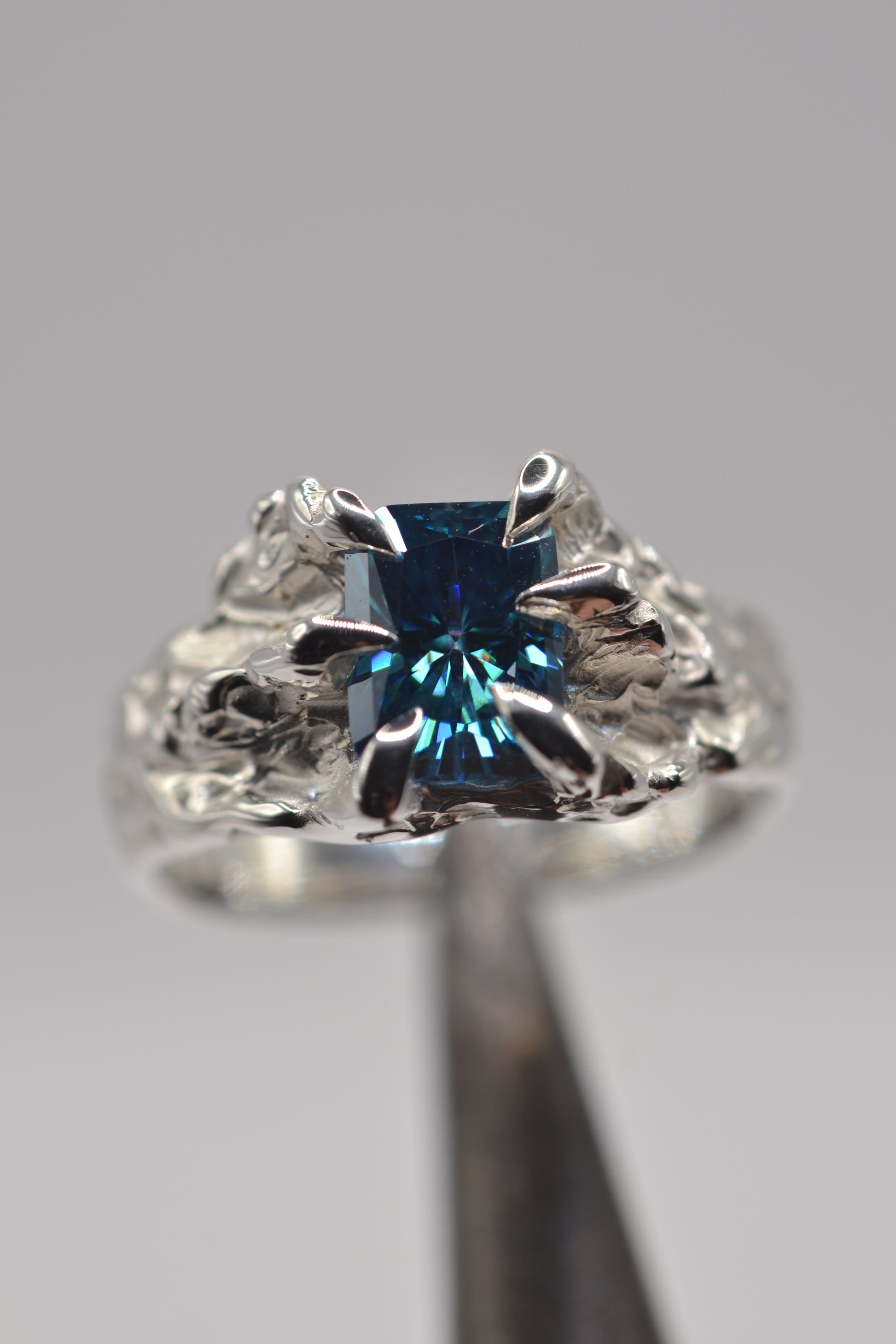 Hero detail of a handmade lost wax silver ring featuring a blue radiant cut moissanite and sculptural organic metal textures.