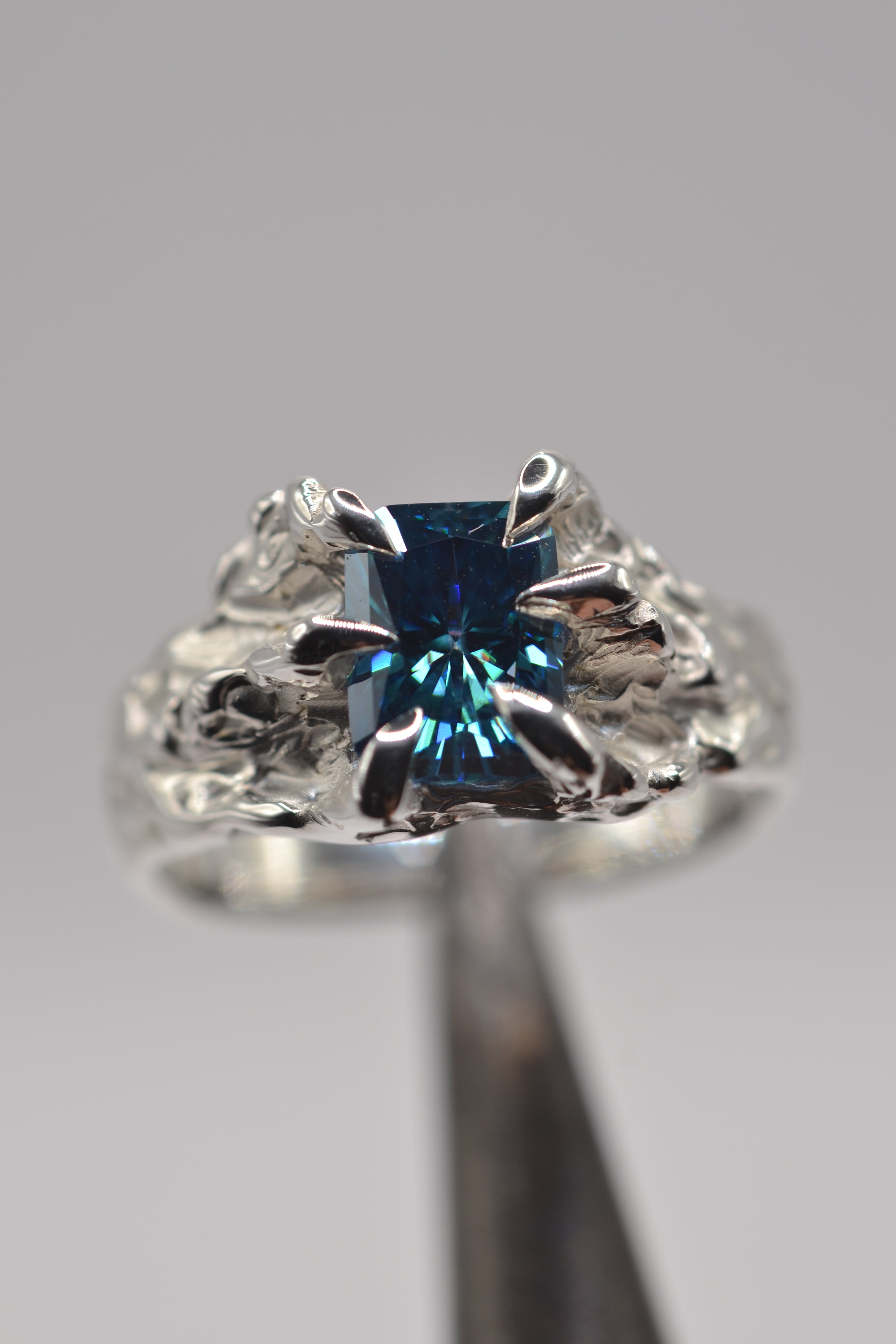 Hero detail of a handmade lost wax silver ring featuring a blue radiant cut moissanite and sculptural organic metal textures.