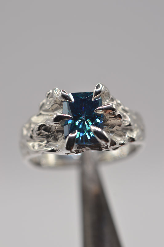 Hero detail of a handmade lost wax silver ring featuring a blue radiant cut moissanite and sculptural organic metal textures.