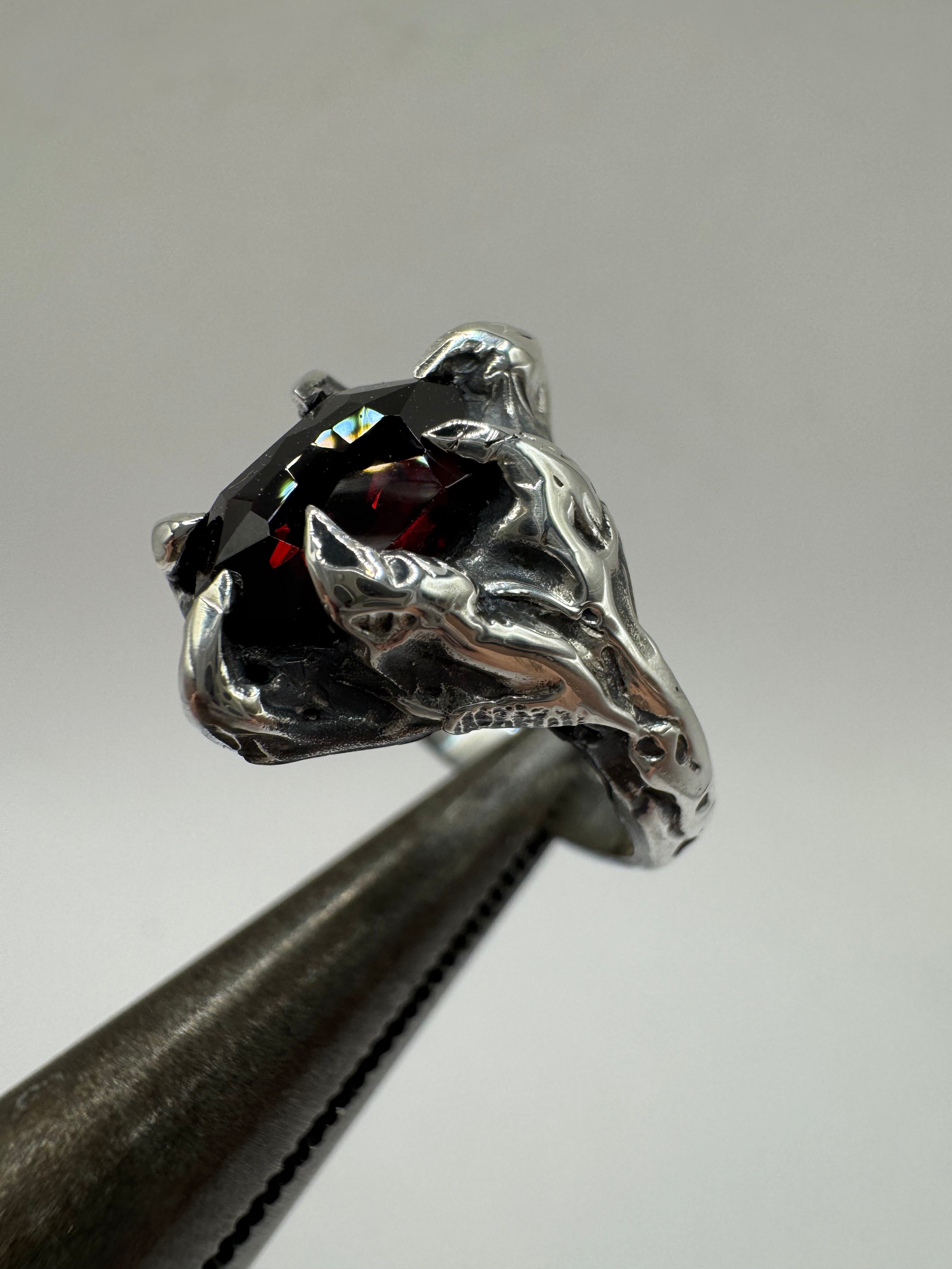 Bold silver ring featuring a deep red faceted garnet set in a six-prong claw setting. Finished with patina and a textured, molten metal look — a perfect alternative jewelry piece.
