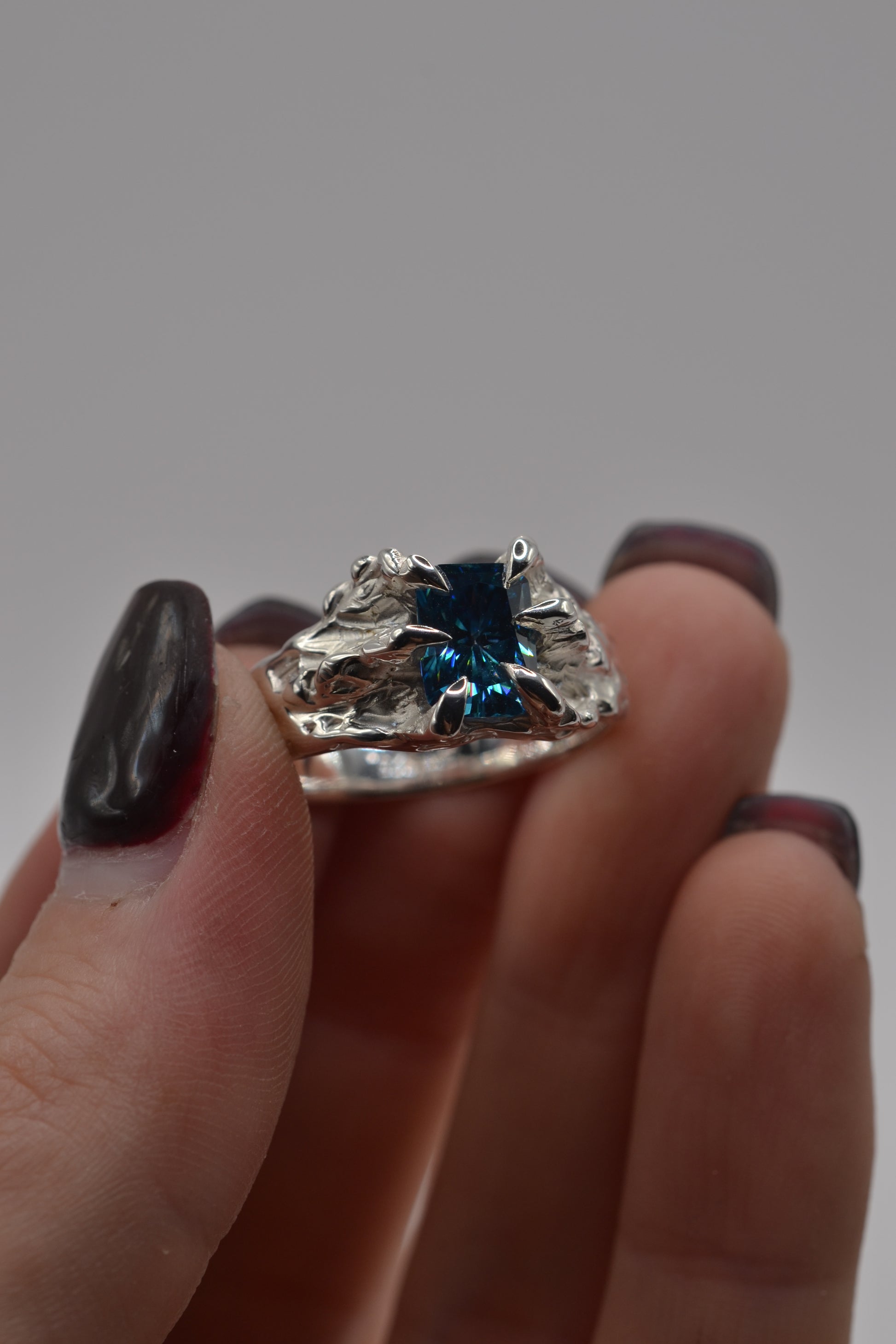 Detailed view of a lost wax cast silver ring with a blue radiant cut moissanite, showcasing organic textures and claw setting.