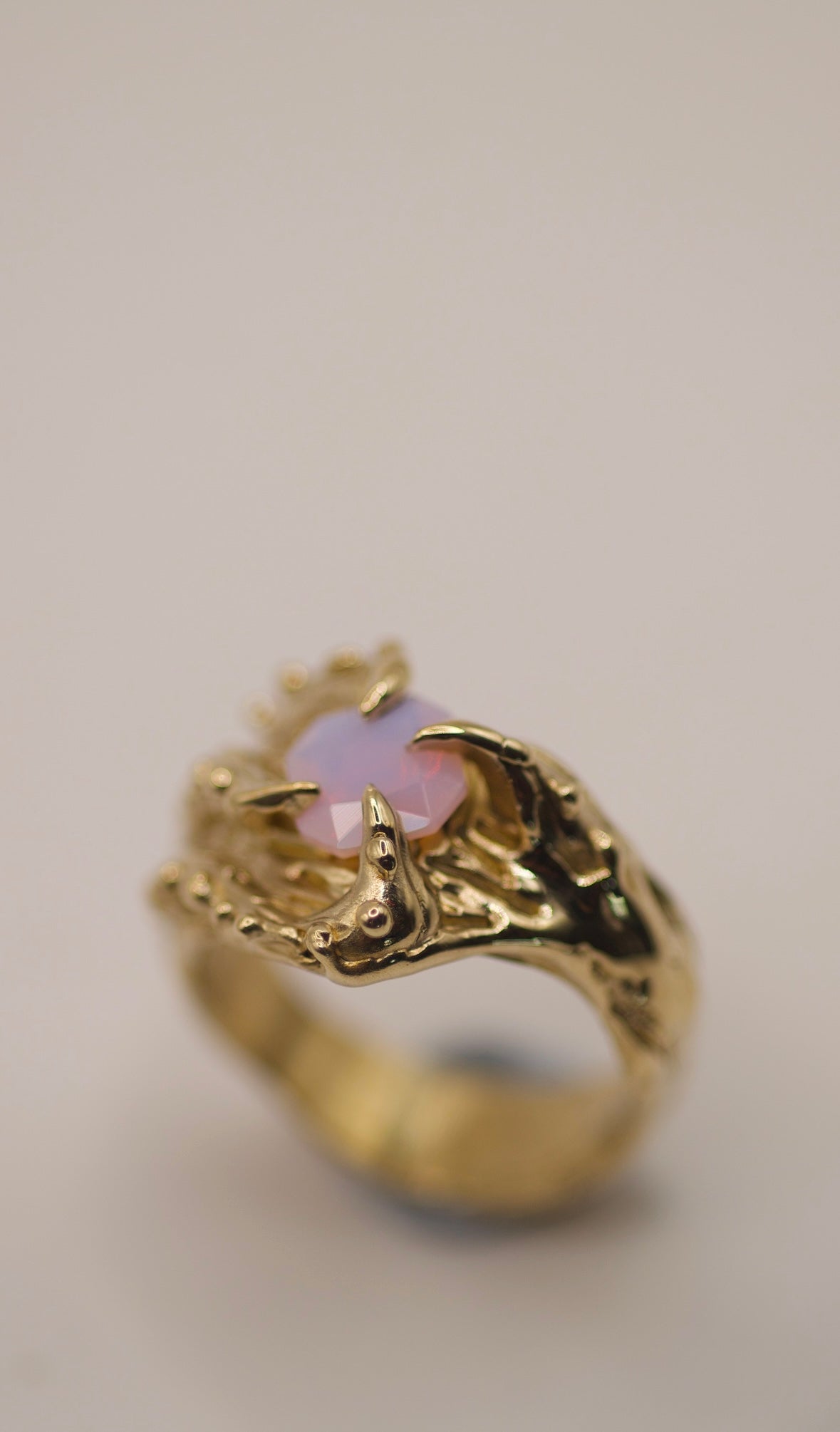 A gold ring that blends organic textures with gothic elements, featuring four claws that curve around a square lab-grown opal with a soft pink glow. The textured band is rich with raised bumps, grooves, and dips, offering a tactile feel reminiscent of an underwater creature or natural form. The opal radiates an ethereal pink hue, contrasting beautifully with the dark, intricate design. This piece is handmade using the lost wax technique, giving it a sculptural, one-of-a-kind quality.