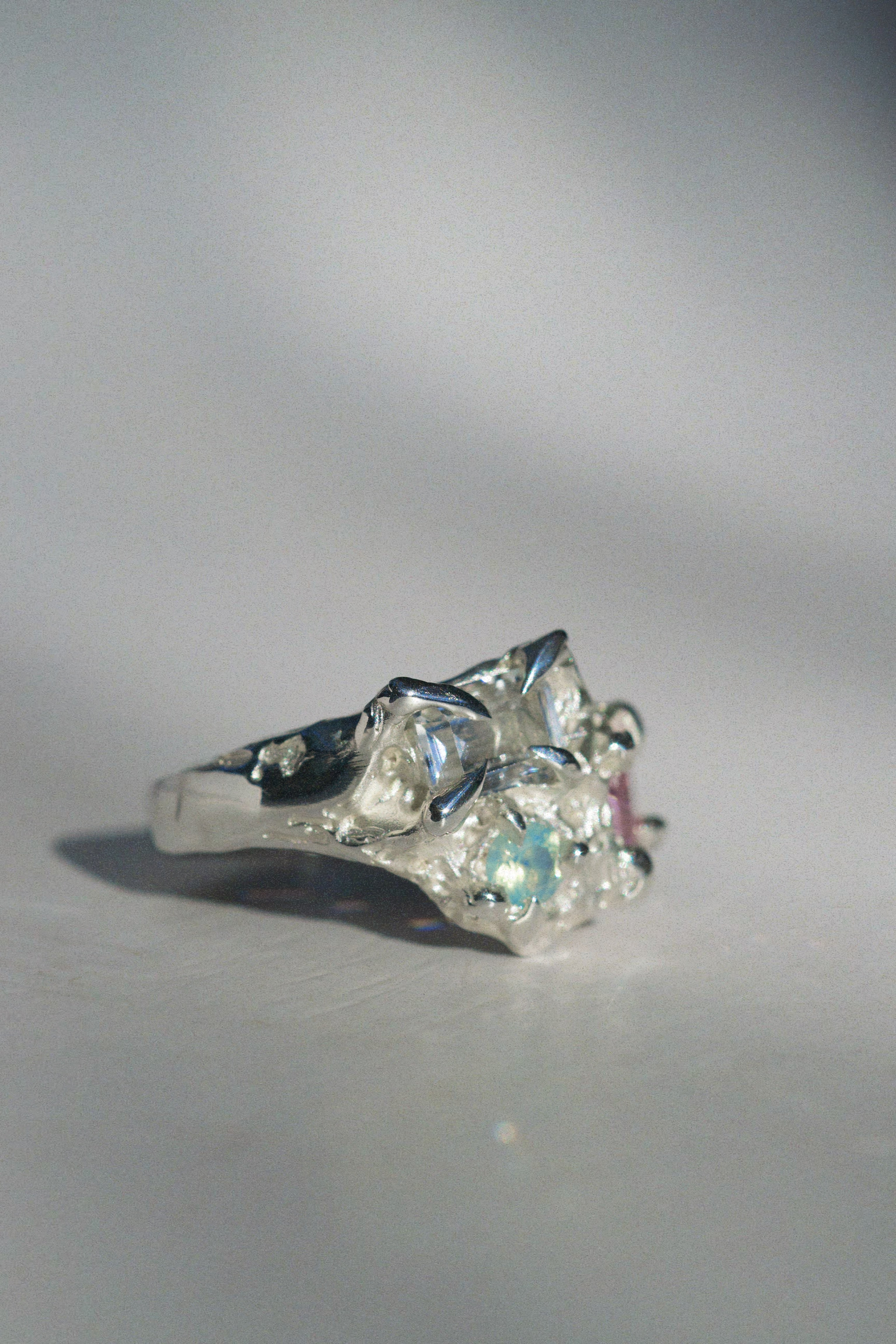 Fantasy-inspired sterling silver ring with three asymmetrical stones — white sapphire, pink CZ, and aqua opal — resting on soft white fabric. The sculptural design mimics blooming petals and molten roots, crafted using the lost wax method.

