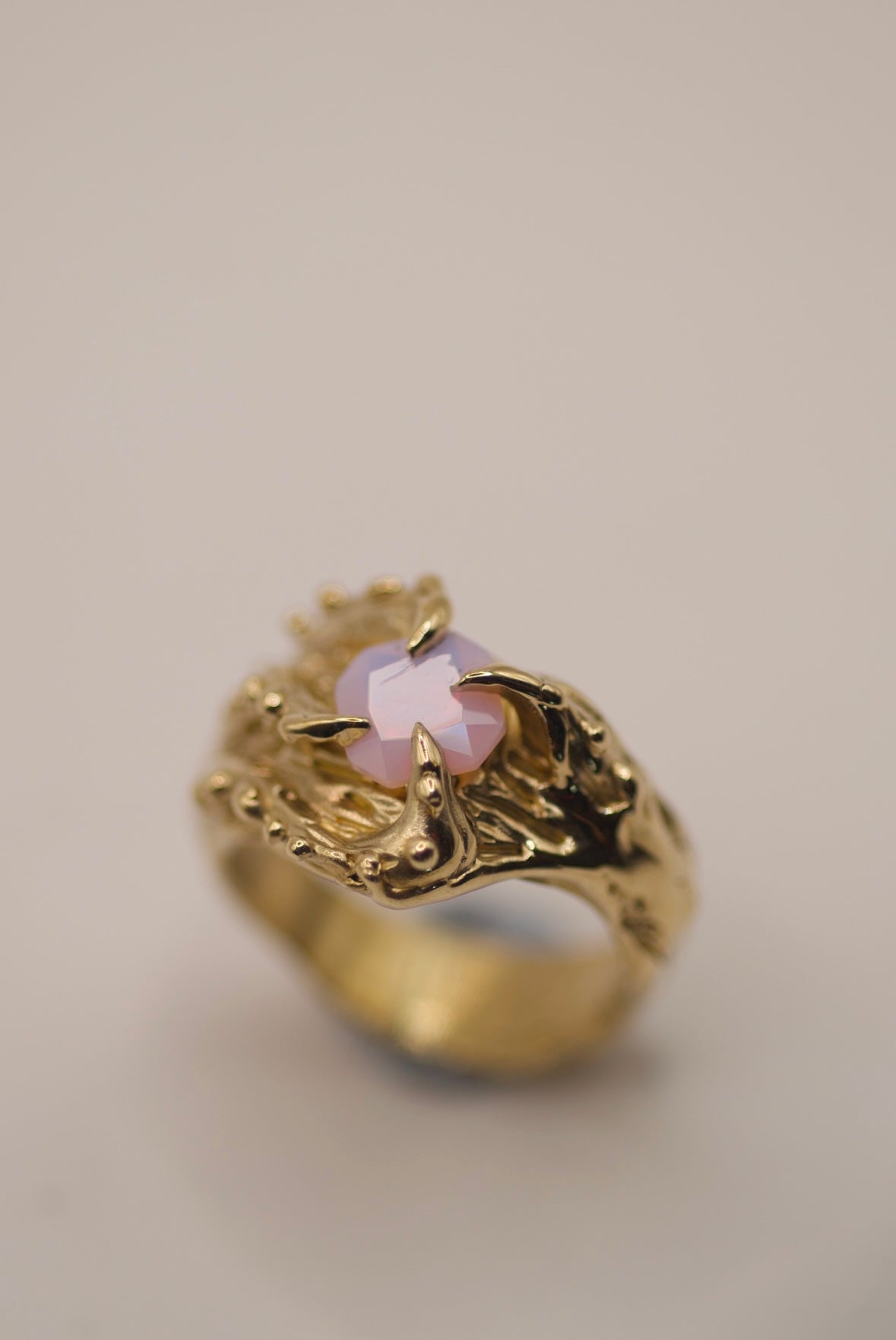 This bold gold ring features four gothic-style curved claws that seem to stretch and grip the square lab-grown opal at its center. The organic texture of the band, full of bumps, dips, and grooves, creates a tactile, fluid design reminiscent of sea creatures or organic forms. The soft pink hue of the opal adds an ethereal glow to the dark, textured gold. Handcrafted with the lost wax technique, this piece has a truly unique, sculptural presence.