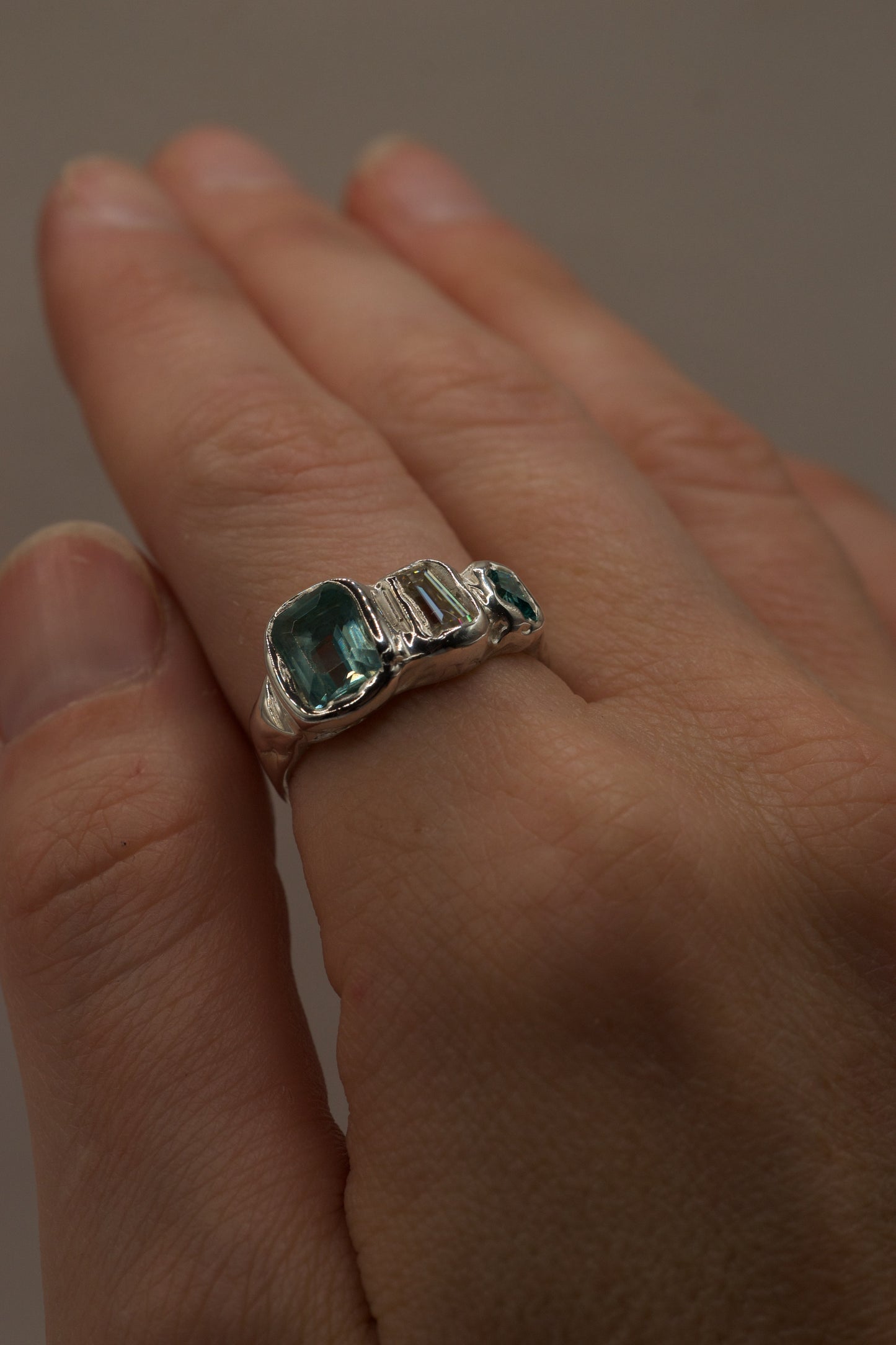 Angled hand-shot of a molten textured silver ring with high polish finish, showcasing a seafoam green gemstone, baguette-cut moissanite, and small green lab-grown crystal, all cast in place through lost wax jewellery making for a bold alternative style.