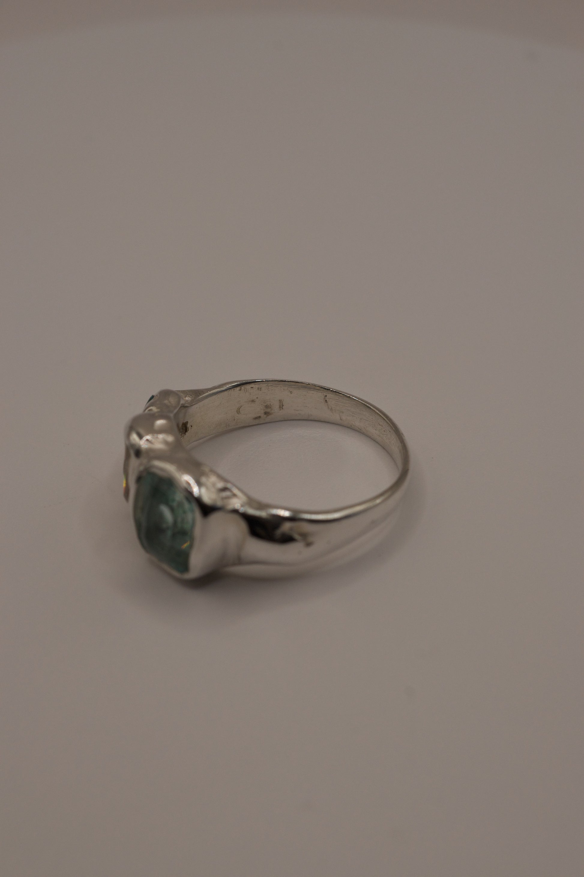 Profile view of an organic silver ring with flowing molten texture, high polish band, and three gemstones cast in place: a central seafoam green crystal, baguette-cut moissanite, and small green crystal, crafted using the lost wax jewellery making process.
