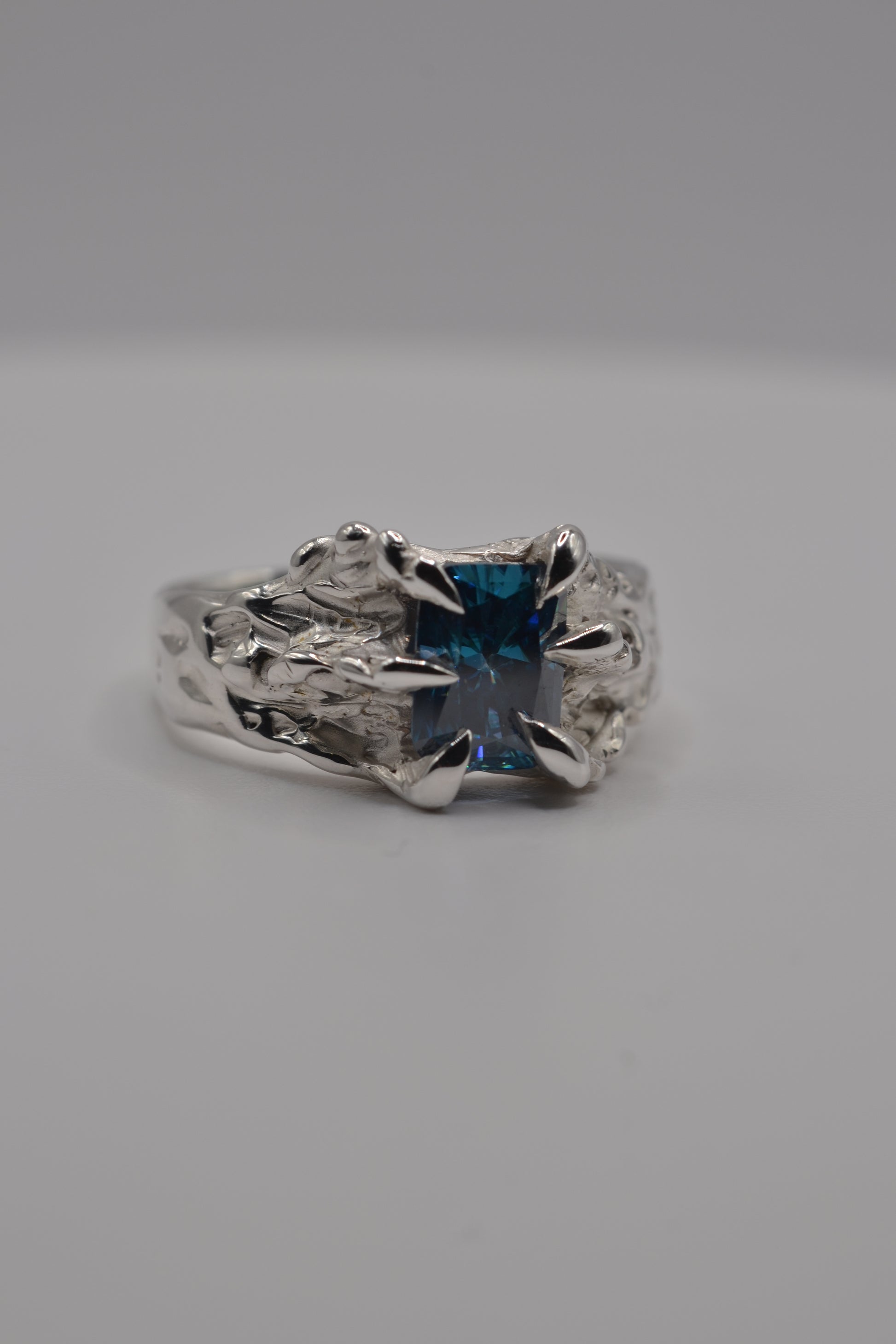 Macro detail of a blue radiant cut moissanite secured in an organic claw setting on a lost wax cast silver ring.