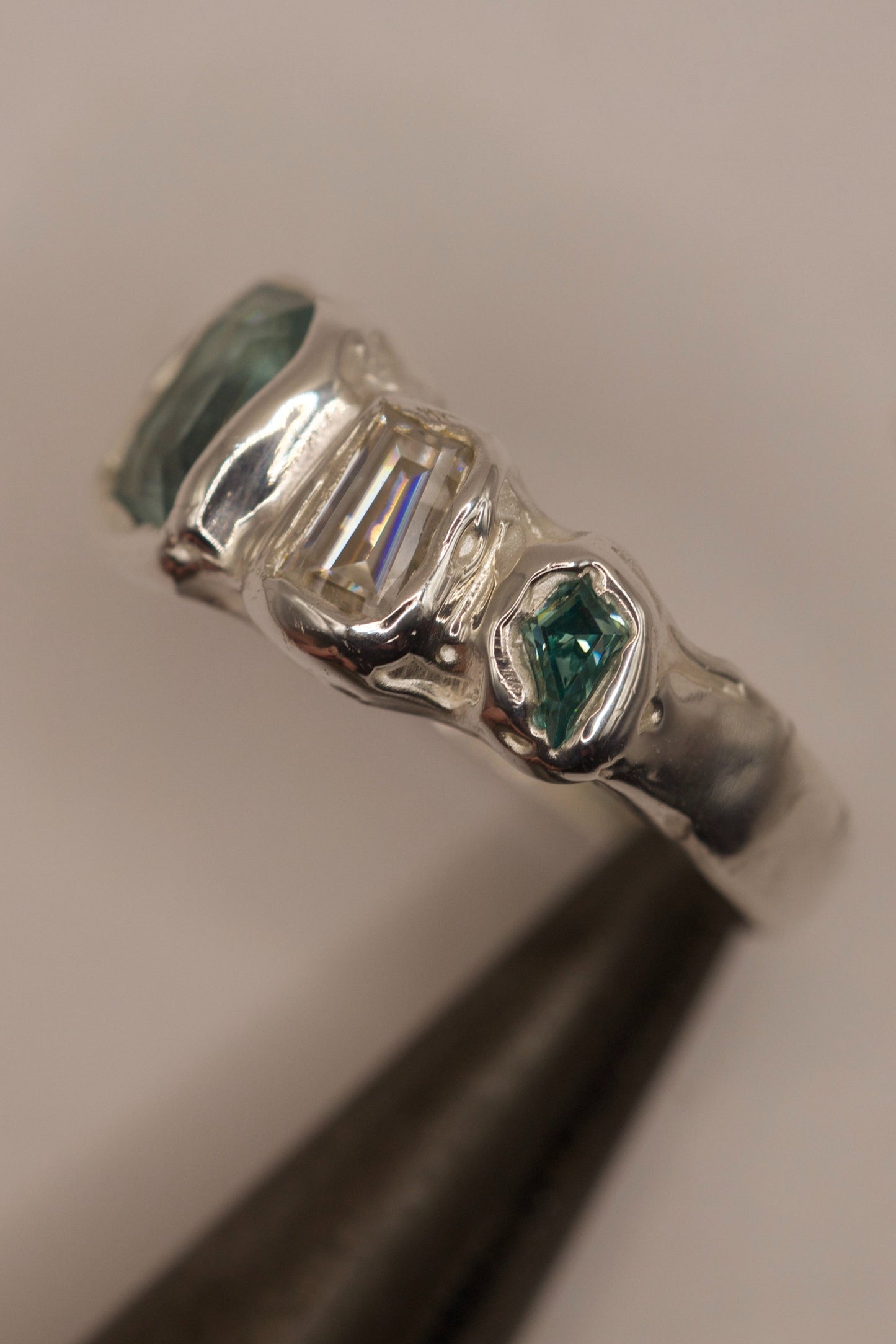 Side view macro of an organic molten silver ring with heavy texture and high polish, featuring a cast in place seafoam green gemstone, baguette moissanite, and a green crystal, handmade for alternative jewellery with the lost wax casting method.