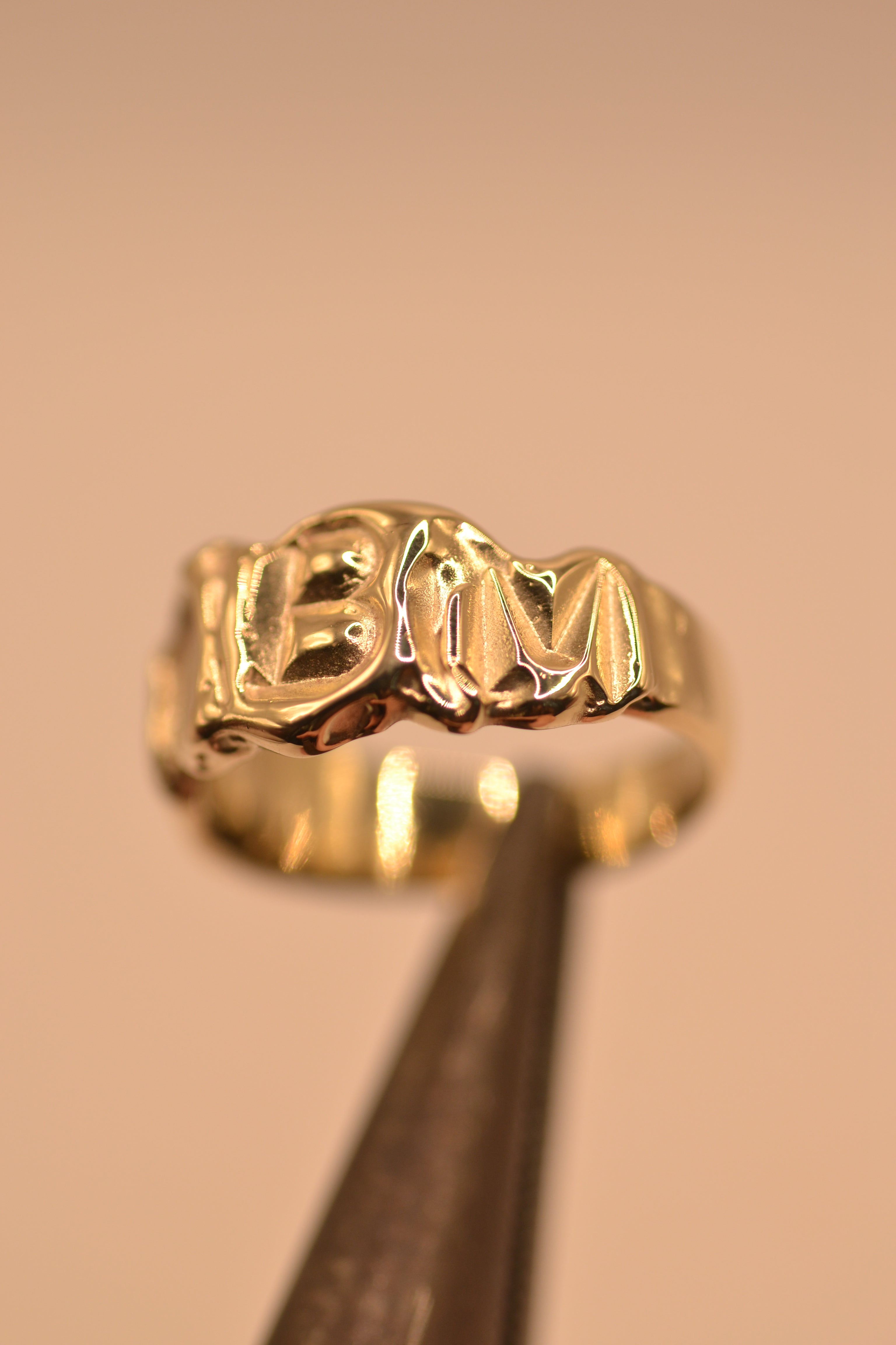 Handmade lost wax cast gold initial ring held in metal tweezers showing molten texture and personalised letters