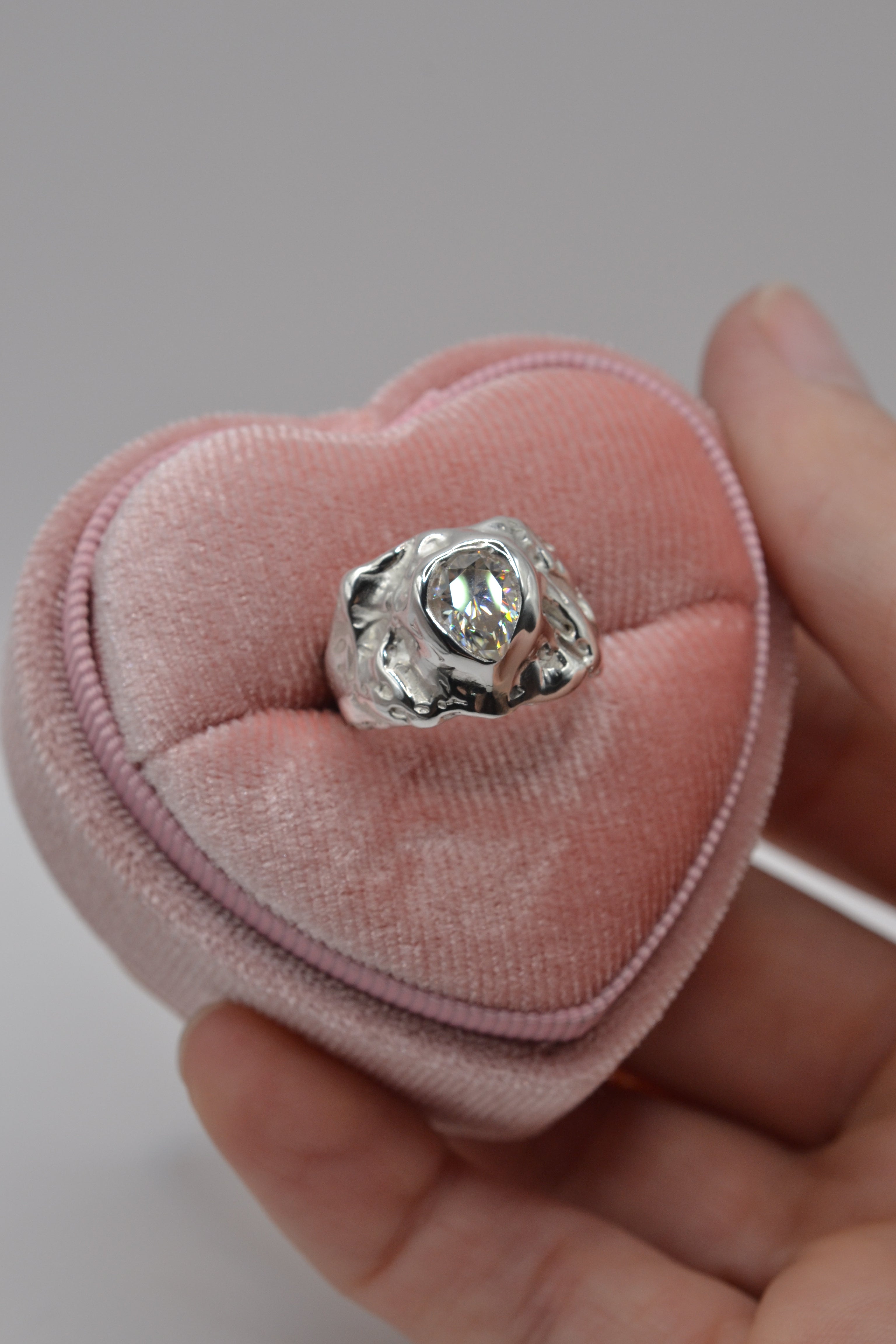 The molten silver lost wax ring with pear-cut moissanite, presented in a pink velvet heart box, showcasing its organic sculptural style.
