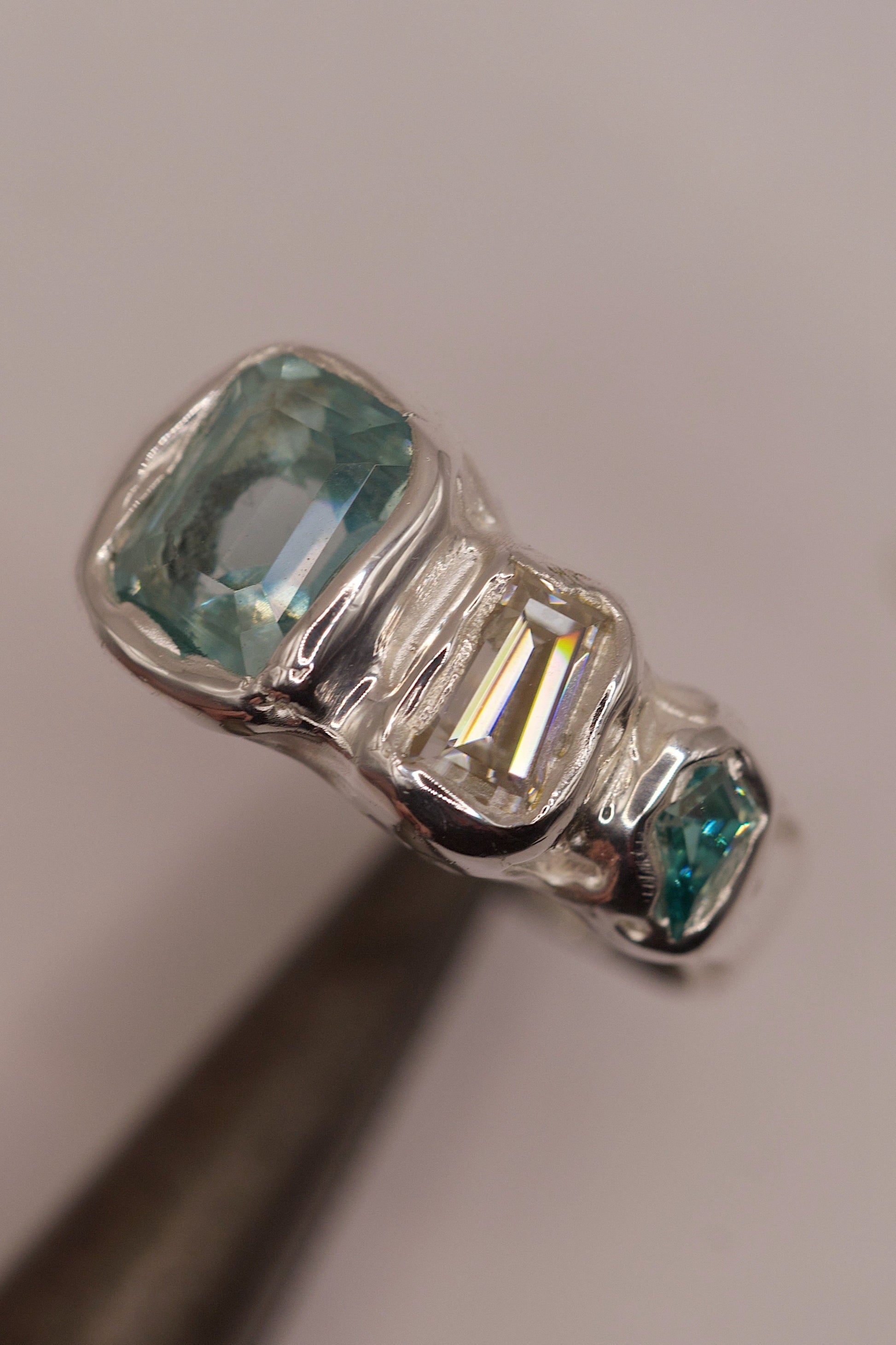 Close-up of a high polish molten 925 sterling silver ring with organic flowing design, displaying three cast in place gemstones: a seafoam green crystal, a baguette-cut sparkling moissanite, and a small green-toned stone, handmade with the lost wax casting process.