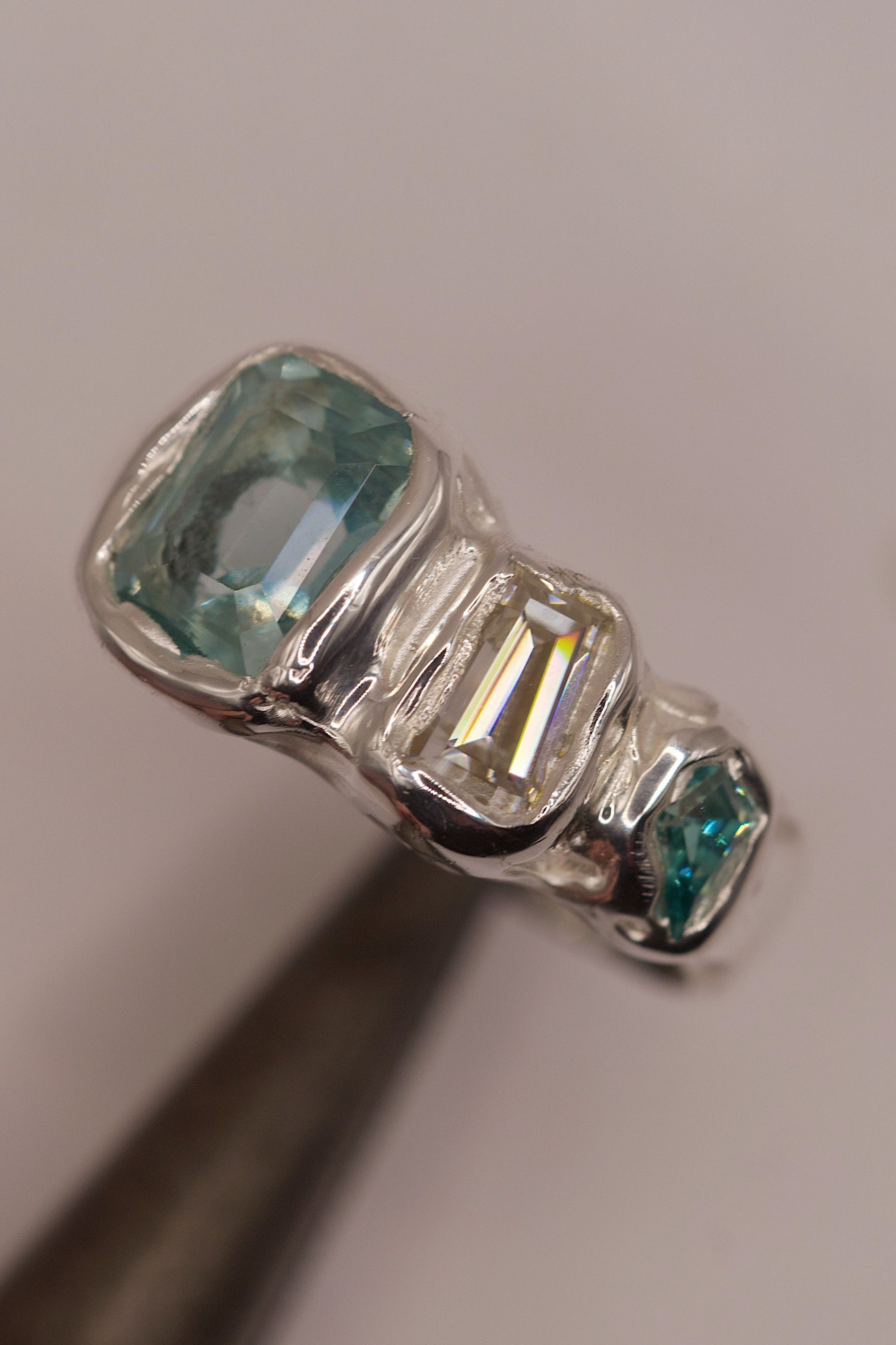 Close-up of a high polish molten 925 sterling silver ring with organic flowing design, displaying three cast in place gemstones: a seafoam green crystal, a baguette-cut sparkling moissanite, and a small green-toned stone, handmade with the lost wax casting process.