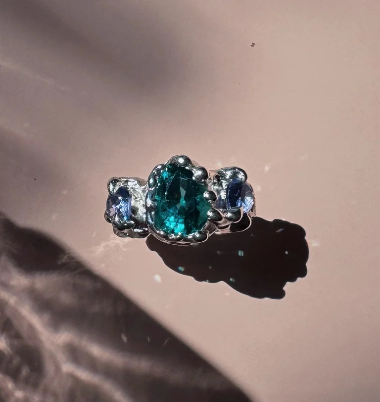 This molten metal sterling silver ring showcases a stunning blue tourmaline at its center, with lilac gemstones adorning the sides. At the heart of the design is a large pear shaped seafoam crystal, beautifully accented by two round amethysts.