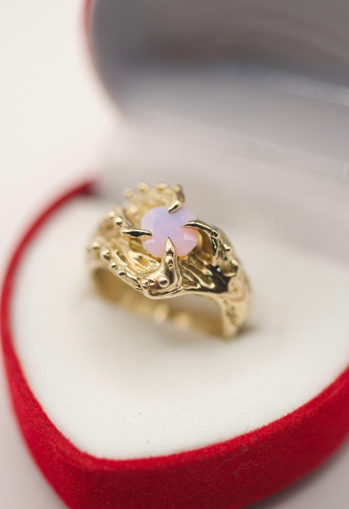 A captivating gold ring with an organic, textured band, featuring four gothic-style curved claws that hold an octagonal-shaped, lab-grown opal. The opal, with its delicate pink hue, sits in stark contrast to the rough, tactile texture of the gold band, which has raised bumps and deep dips. Handmade with precision using the lost wax technique, this ring combines natural elements with gothic elegance, creating a piece that feels both timeless and otherworldly.