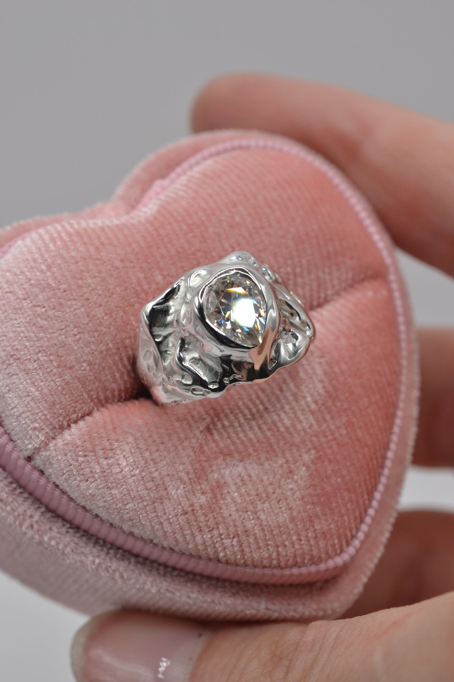 An angled view of the molten silver moissanite ring showcasing bold textures and sculpted lost wax design on a pink heart box.