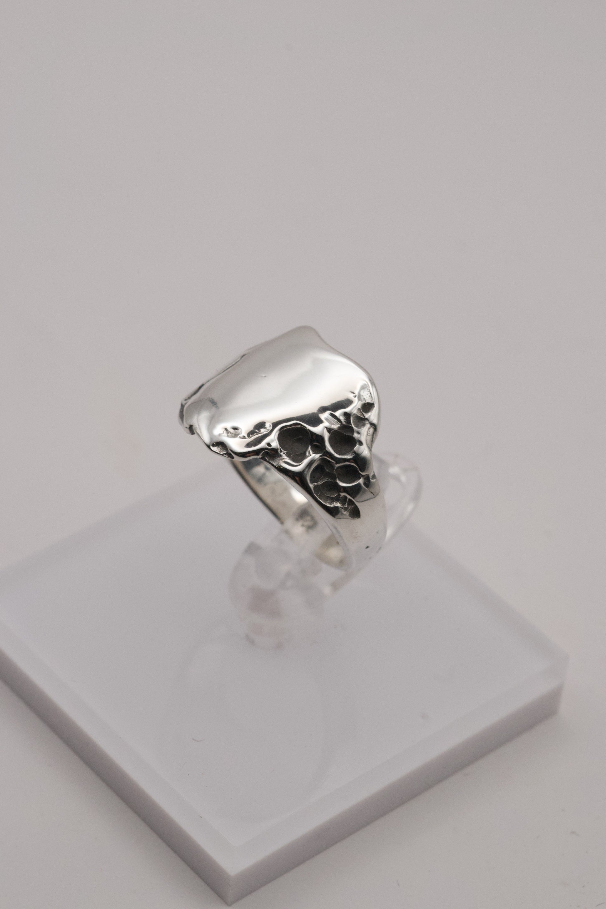 A handcrafted sterling silver signet ring displayed on a white stand, featuring a smooth top and rugged molten edges sculpted in wax.