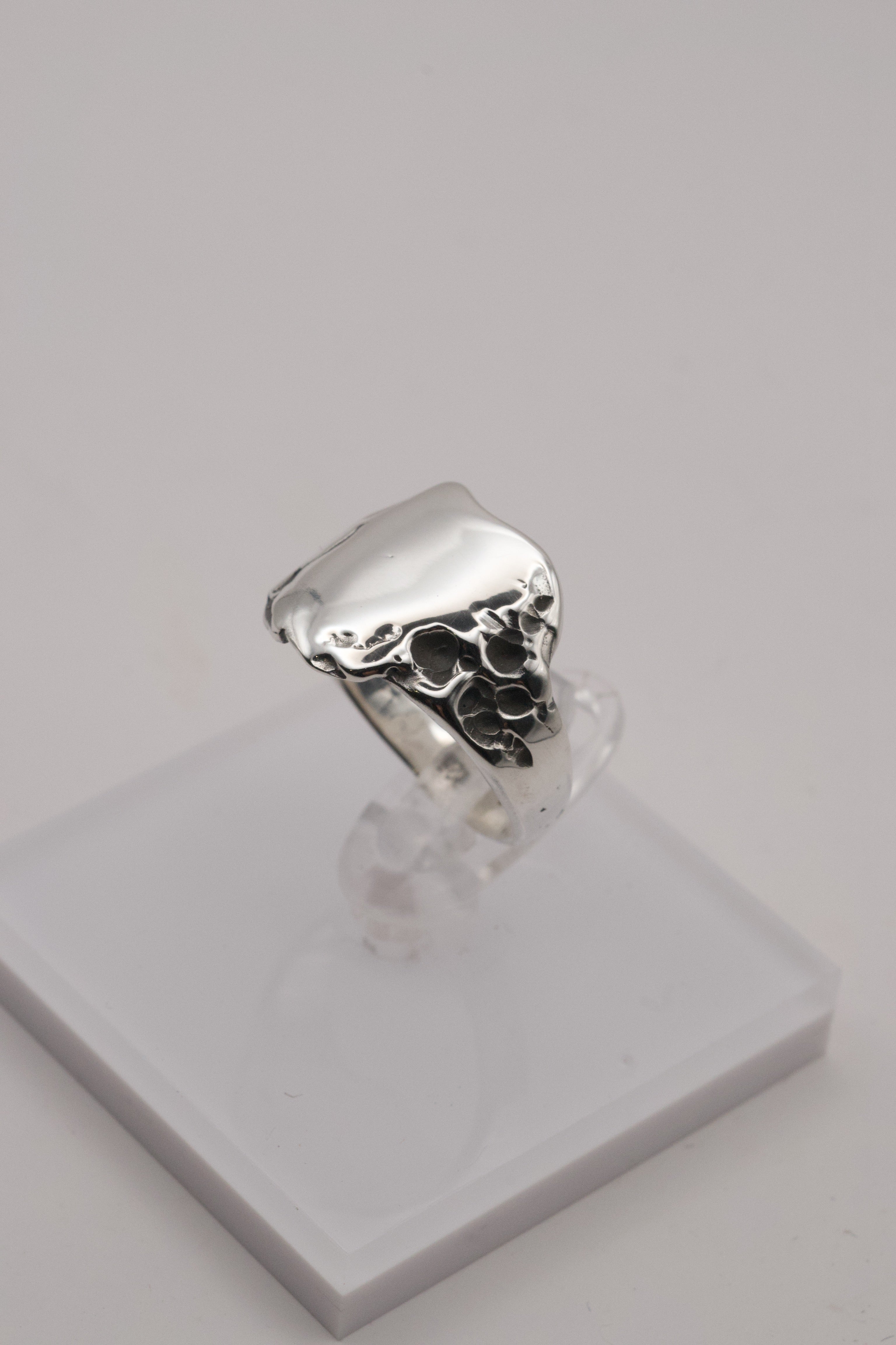 A handcrafted sterling silver signet ring displayed on a white stand, featuring a smooth top and rugged molten edges sculpted in wax.