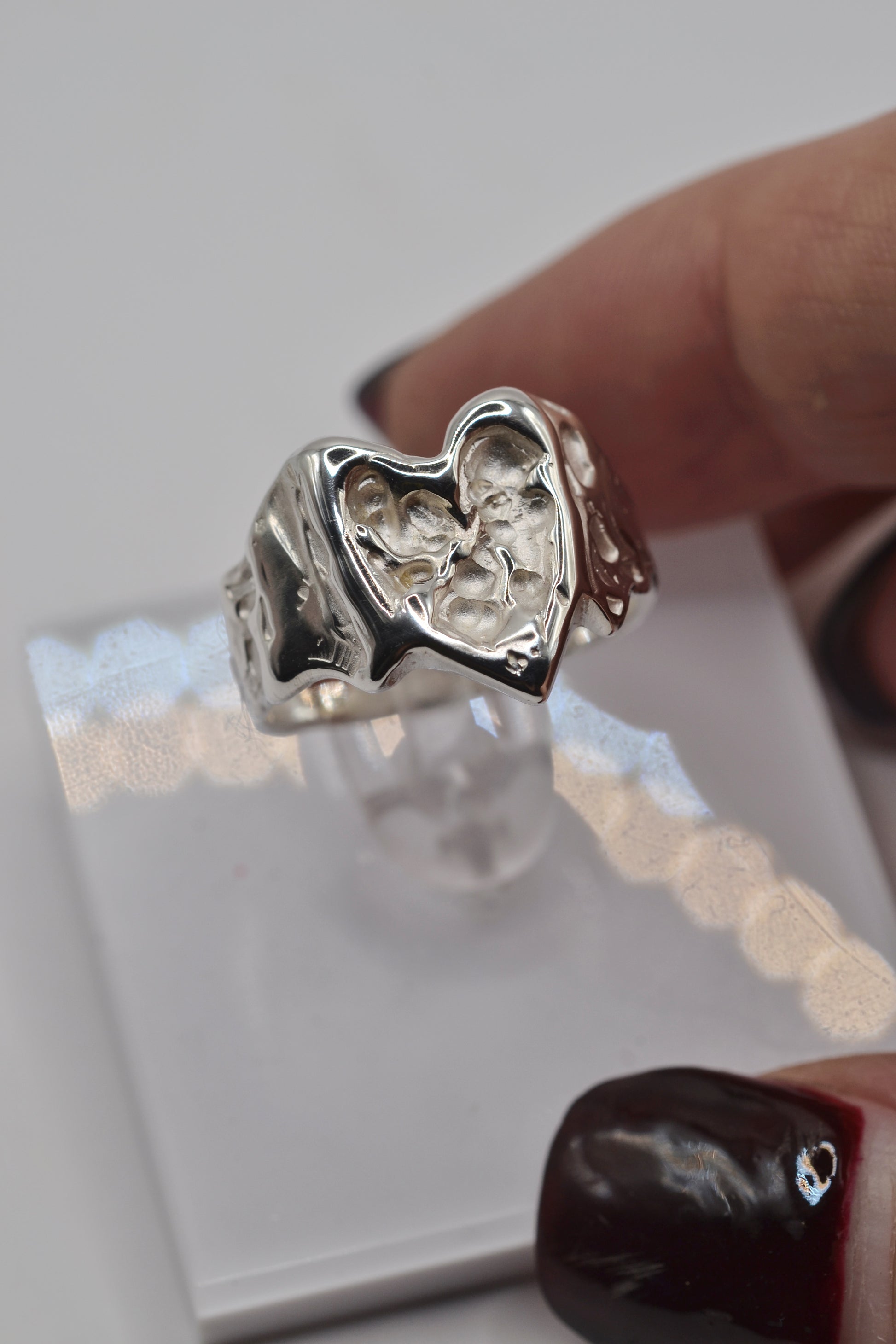 Organic lost wax cast ring in sterling silver with molten heart design, photographed on clear stand and held between fingers, handmade alternative jewellery