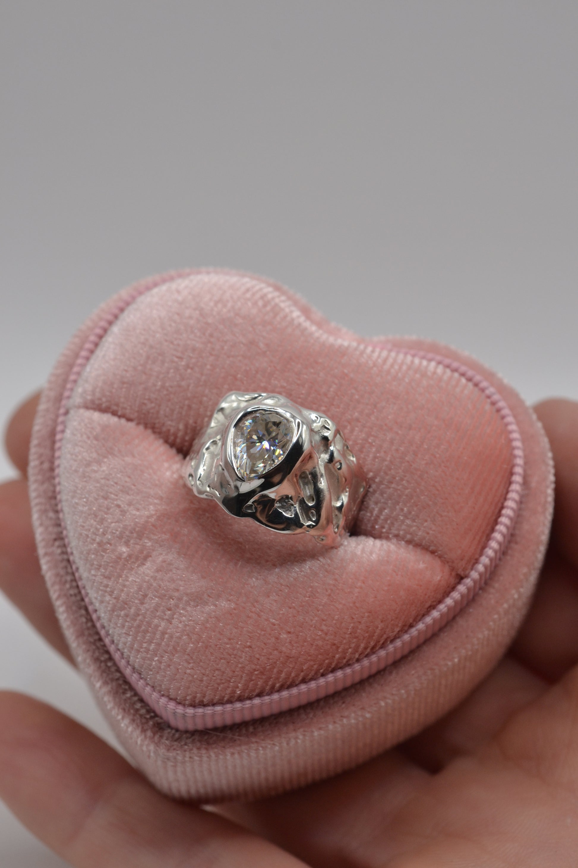 A molten silver pear-cut moissanite ring resting in a velvet heart ring box, highlighting its handmade lost wax detailing.