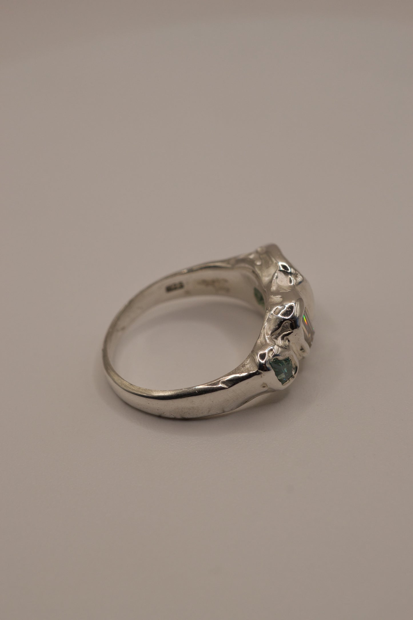 Side angle of an alternative organic sterling silver ring with smooth high polish finish and molten texture, displaying cast in place gemstones including a square seafoam green stone, a baguette-cut moissanite, and a green lab-grown crystal, handmade via lost wax casting.