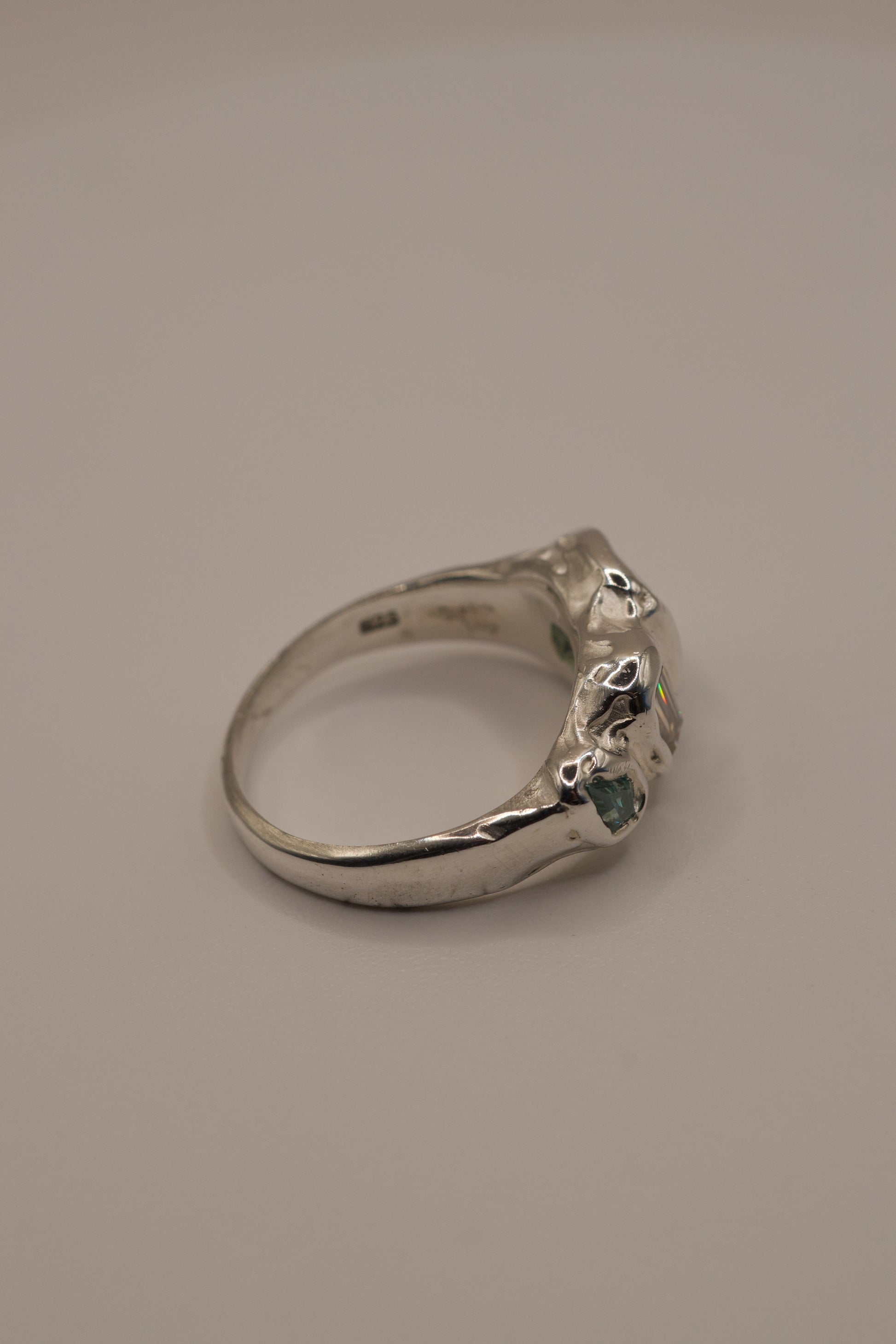 Side angle of an alternative organic sterling silver ring with smooth high polish finish and molten texture, displaying cast in place gemstones including a square seafoam green stone, a baguette-cut moissanite, and a green lab-grown crystal, handmade via lost wax casting.