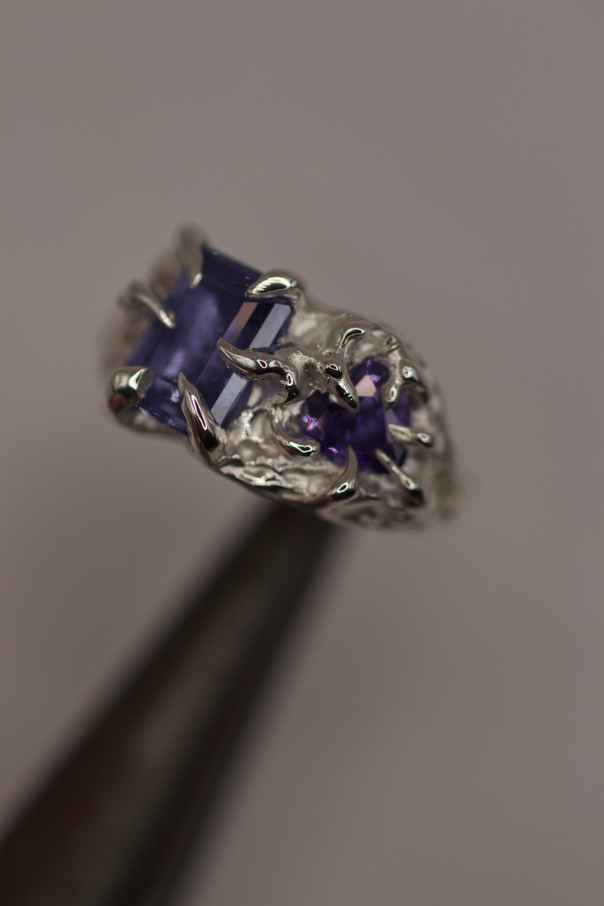 Close-up of a handmade silver ring with a deep purple rectangular gemstone held in a gothic-style claw setting. The design is textured and organic, created using the lost wax casting method.