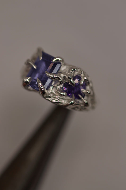 Close-up of a handmade silver ring with a deep purple rectangular gemstone held in a gothic-style claw setting. The design is textured and organic, created using the lost wax casting method.