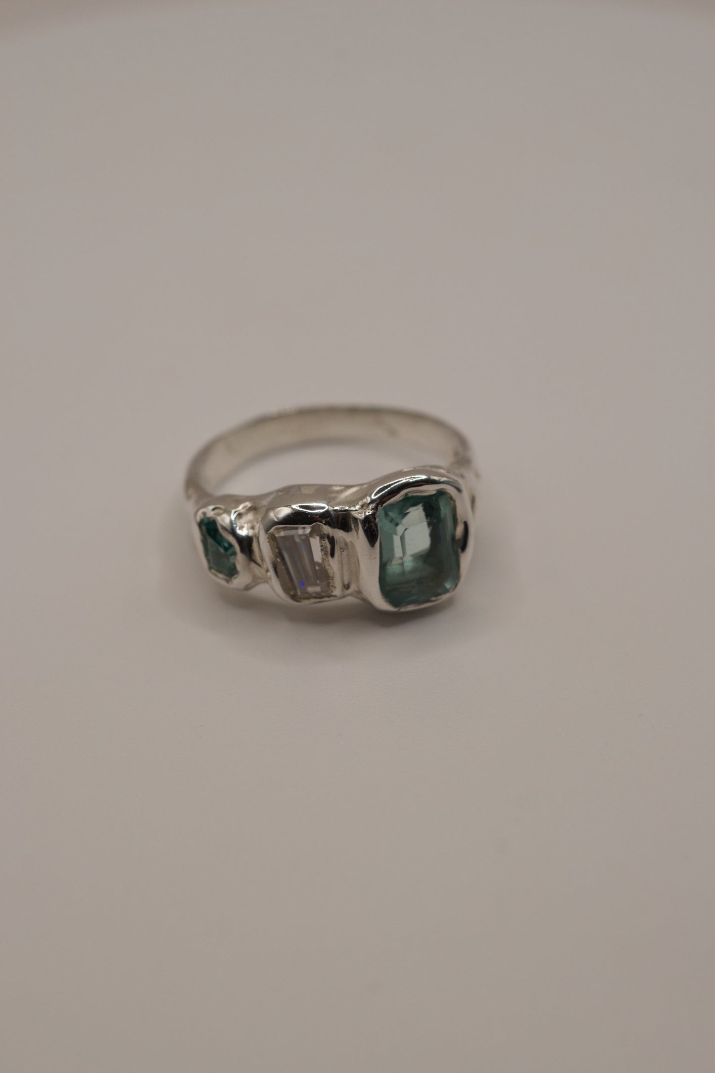 Highly textured sterling silver ring in an organic molten style, high polish metal showcasing three gemstones: a seafoam green lab-grown crystal, a baguette-cut moissanite, and a smaller green crystal, all set with cast in place technique through lost wax jewellery making.