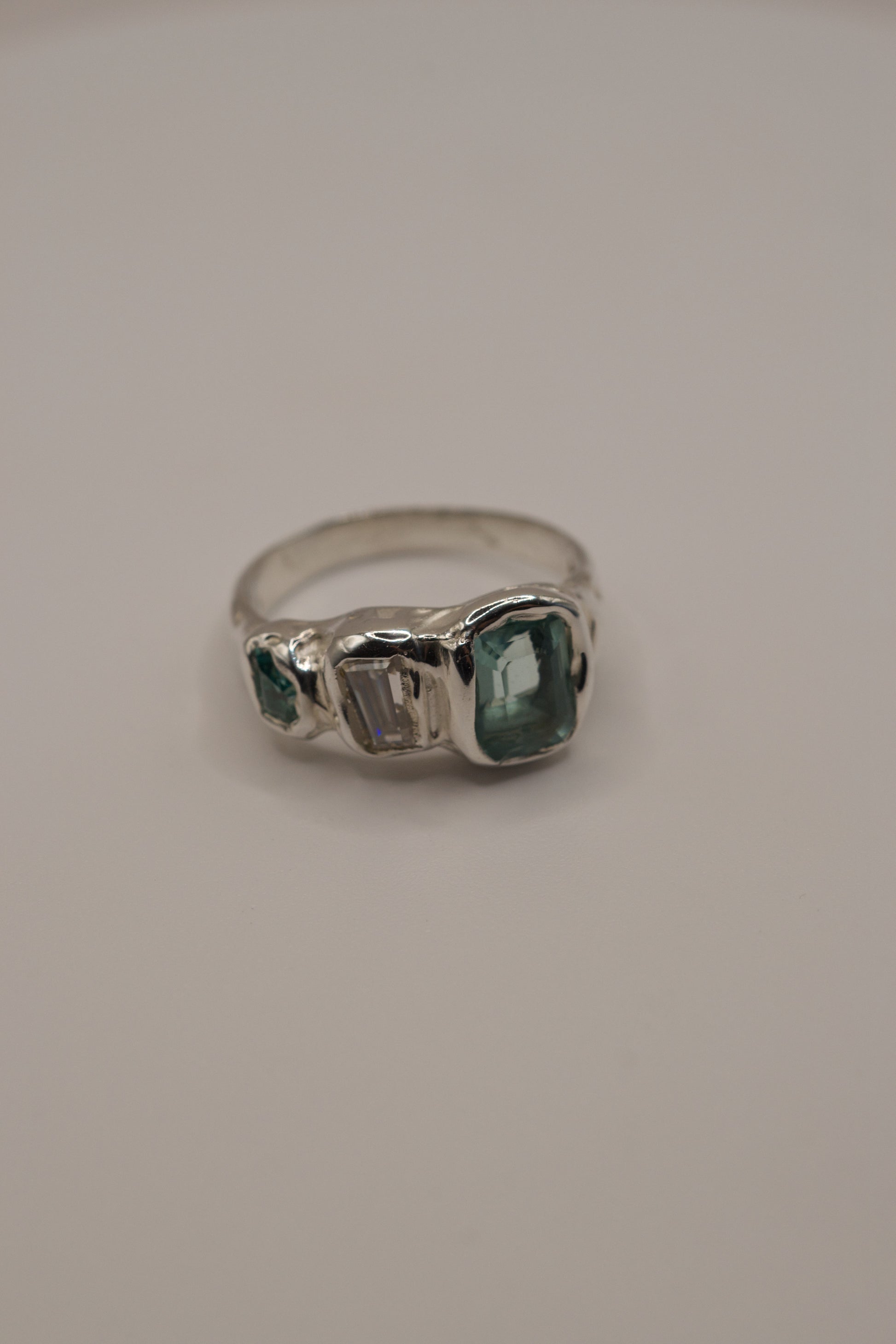 Highly textured sterling silver ring in an organic molten style, high polish metal showcasing three gemstones: a seafoam green lab-grown crystal, a baguette-cut moissanite, and a smaller green crystal, all set with cast in place technique through lost wax jewellery making.