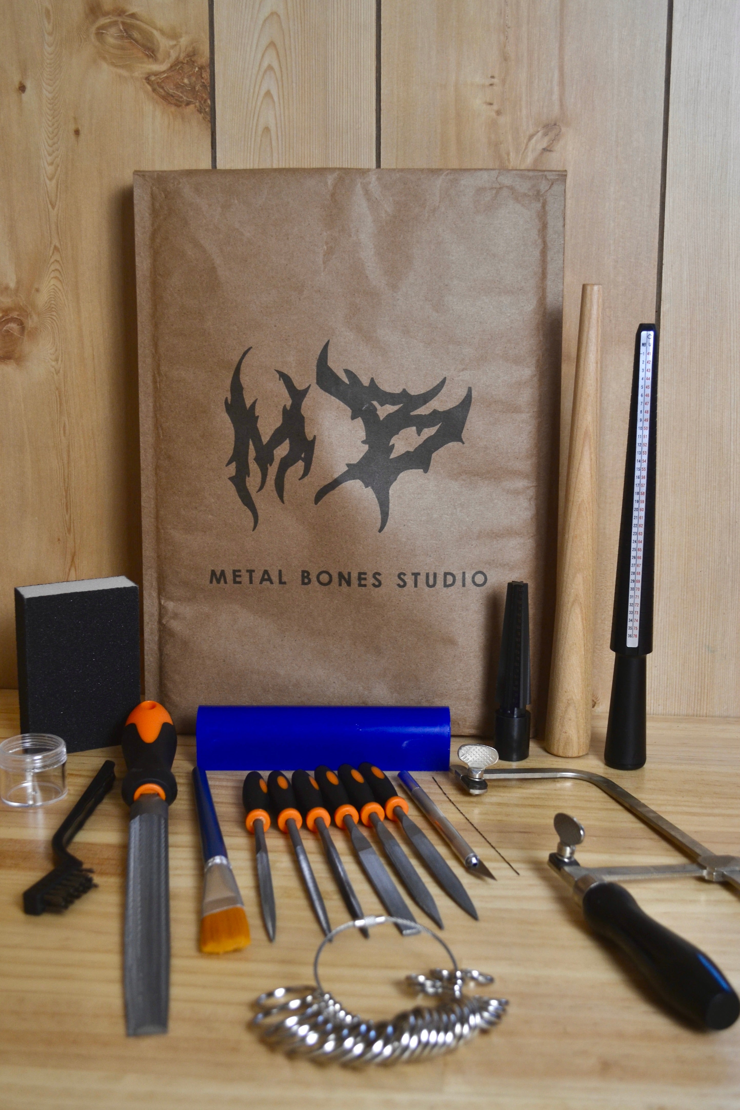 A beginner friendly wax carving kit from Metal Bones Studio arranged on a wooden surface in front of a brown paper bag with the Metal Bones logo. The kit includes various jewellery making tools such as files, a wood mandrel, a mini wax carving mandrel, a craft knife, and a small brass wire brush, alongside genuine blue ferris wax.