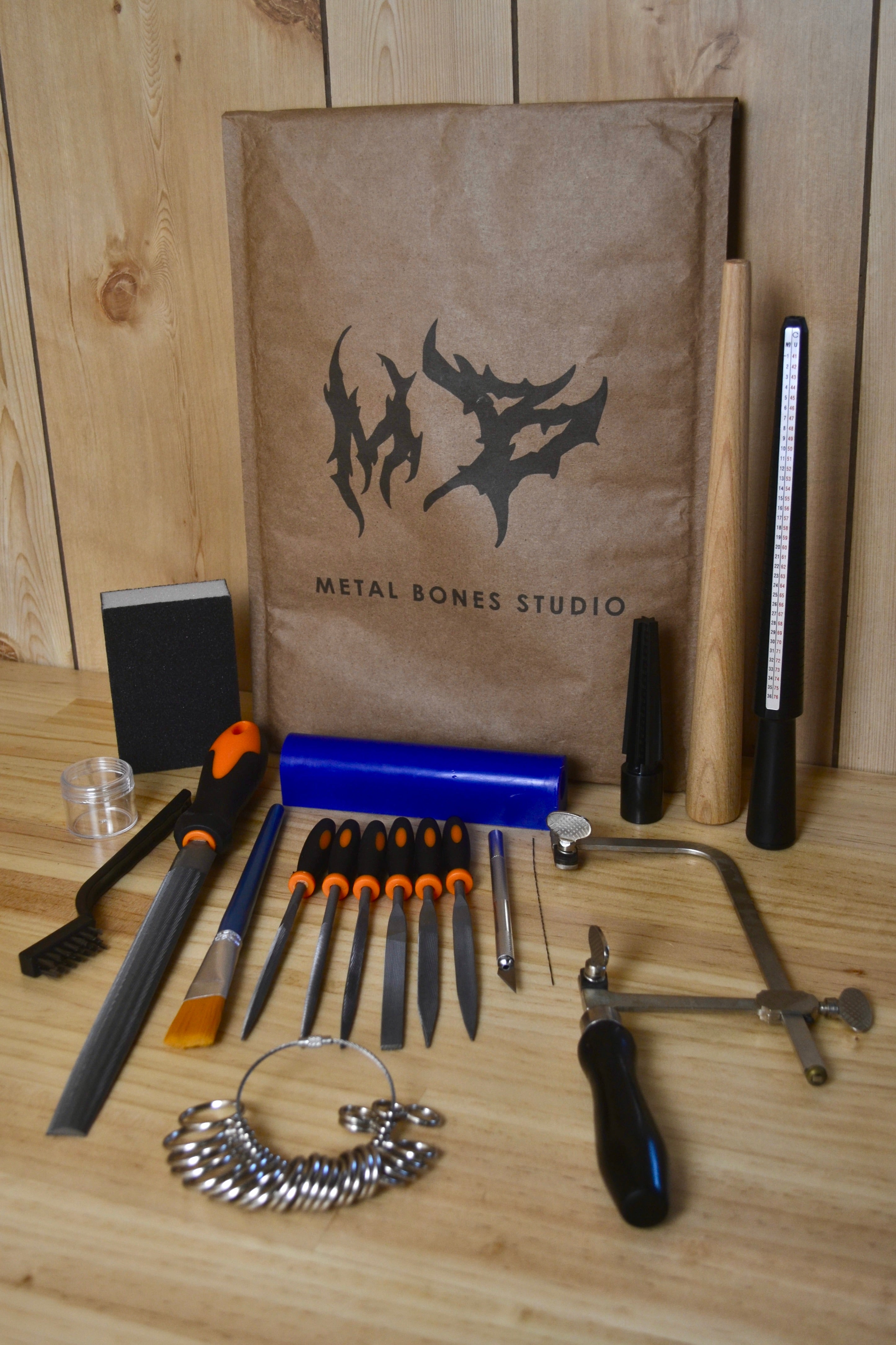 A beginner friendly wax carving kit from Metal Bones Studio arranged on a wooden surface in front of a brown paper bag with the Metal Bones logo. The kit includes various jewellery making tools such as files, a wood mandrel, a mini wax carving mandrel, a craft knife, and a small brass wire brush, alongside genuine blue ferris wax.