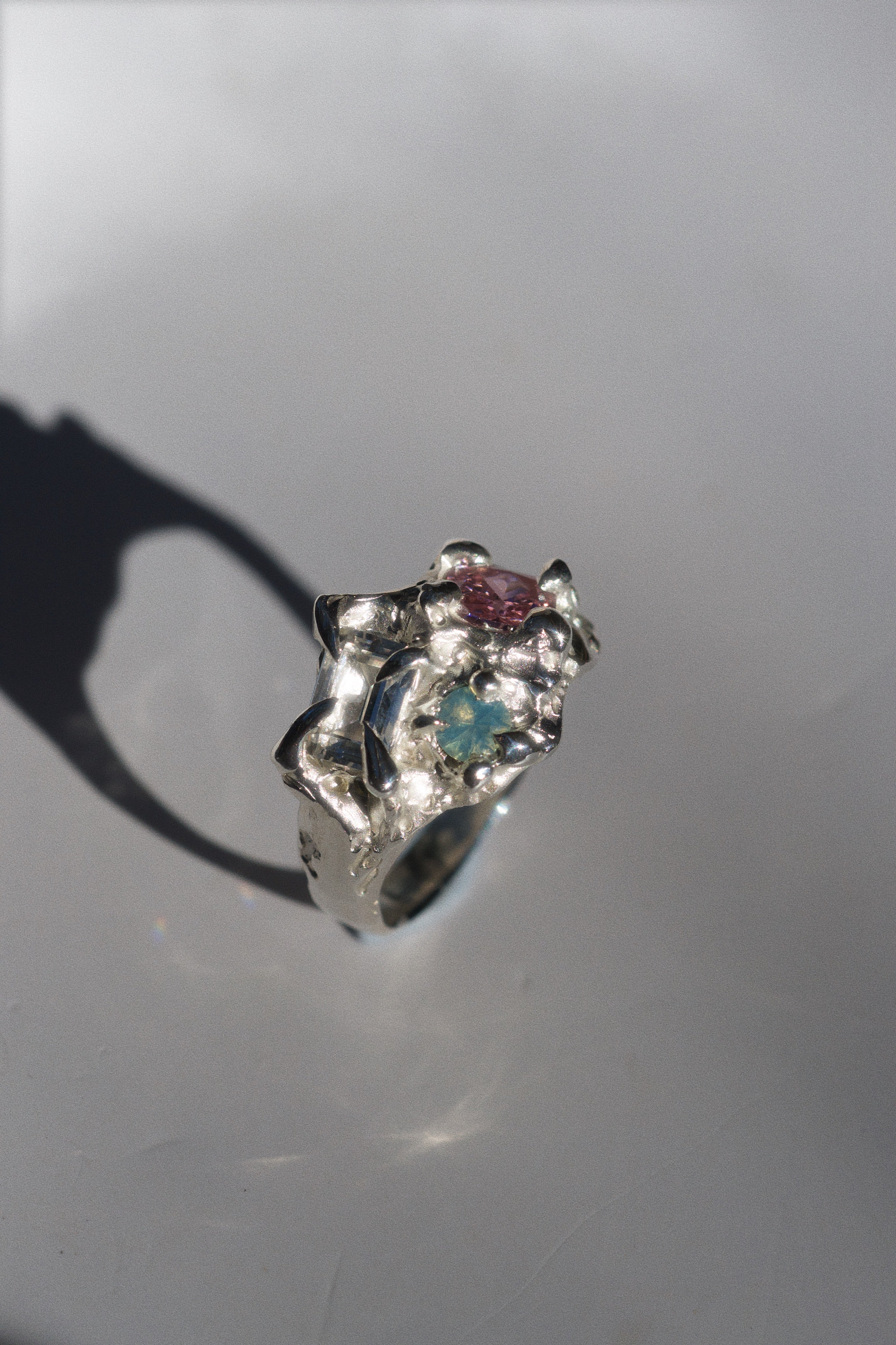 Close-up of gemstone settings on a fantasy-inspired silver ring: emerald-cut white sapphire, cushion-cut pink CZ, and round aqua opal. The textured silver flows around them like melted vines or twisted roots.