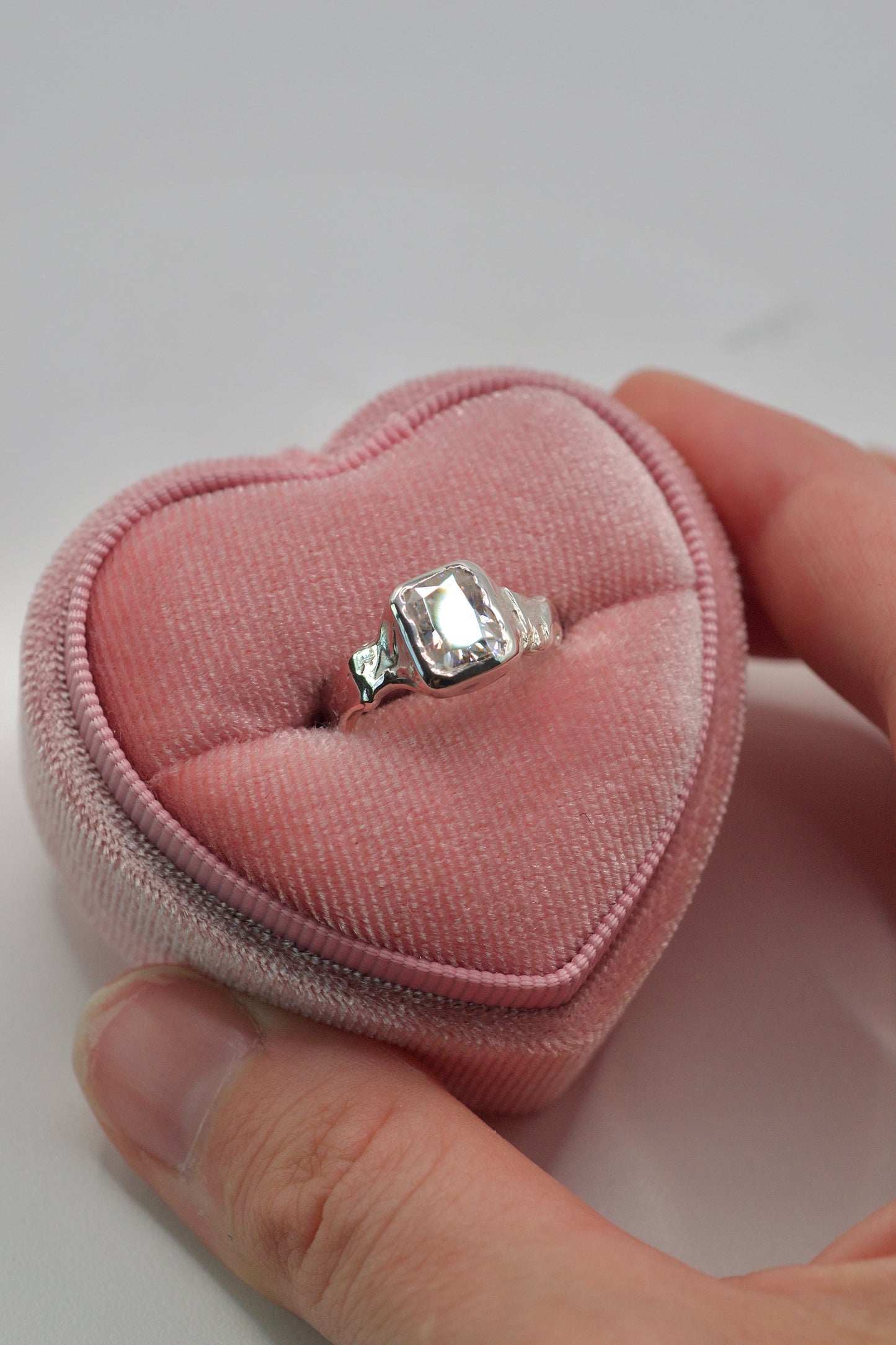 hand holding a sterling silver organic ring with radiant cut white moissanite in bezel setting sitting in a pink velvet heart ring box