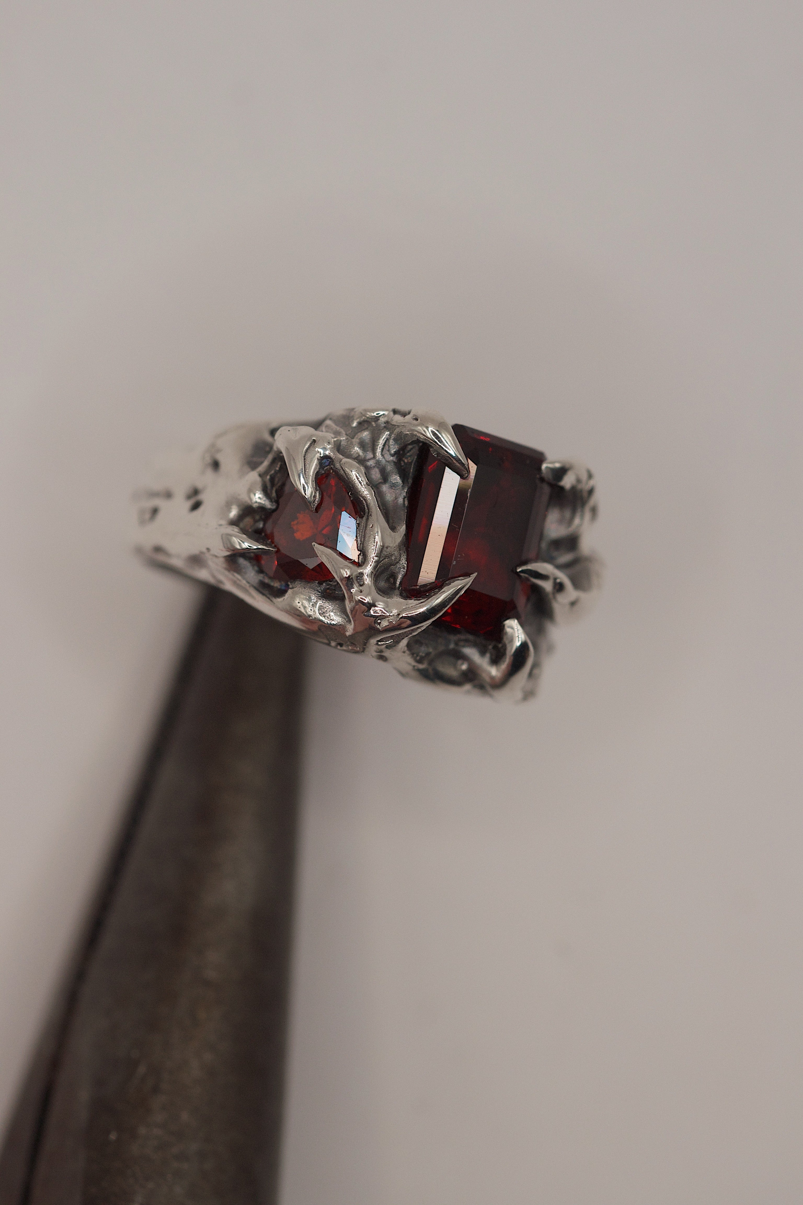 Cherry Ring held in tweezers showing red lab-grown garnet and heart cubic zirconia within sharp organic sterling silver claws handmade using the lost wax technique