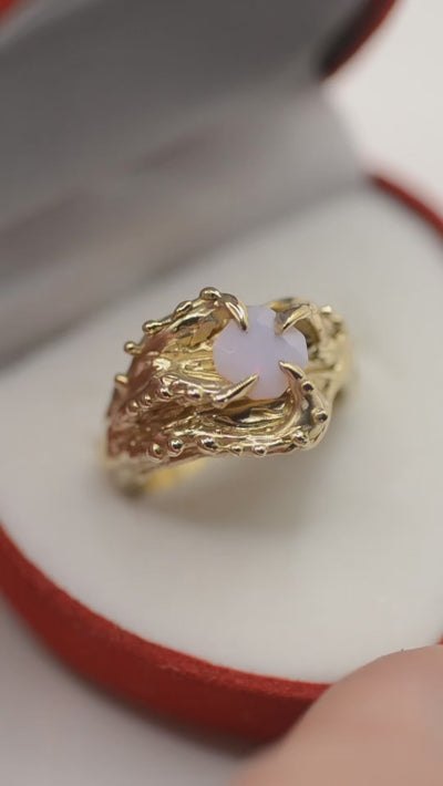 A unique, handmade gold ring with an organic, textured design. The band has gothic, octopus-like features, with four curved claws that wrap around the centerpiece. The ring's surface is full of raised bumps, dips, and grooves, creating a tactile, intricate pattern. In the center, a square-shaped lab-grown opal with a soft pink hue sits, glowing with an ethereal, almost otherworldly light. This sculptural piece was crafted using the lost wax technique, giving it a one-of-a-kind, handcrafted look video.
