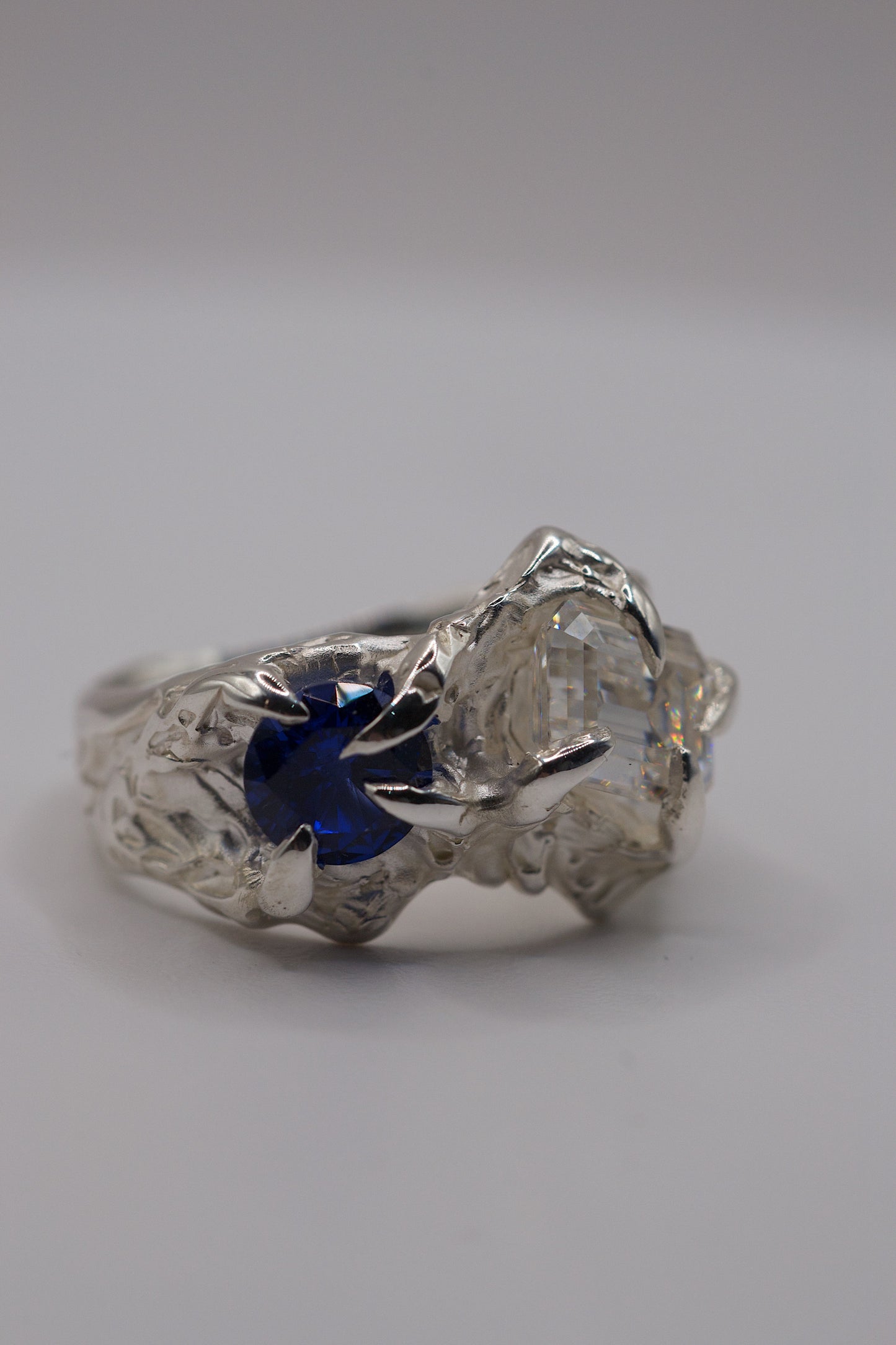 Handmade sterling silver alternative lost wax ring with round blue sapphire and emerald cut moissanite held in place by sharp gothic claws.
