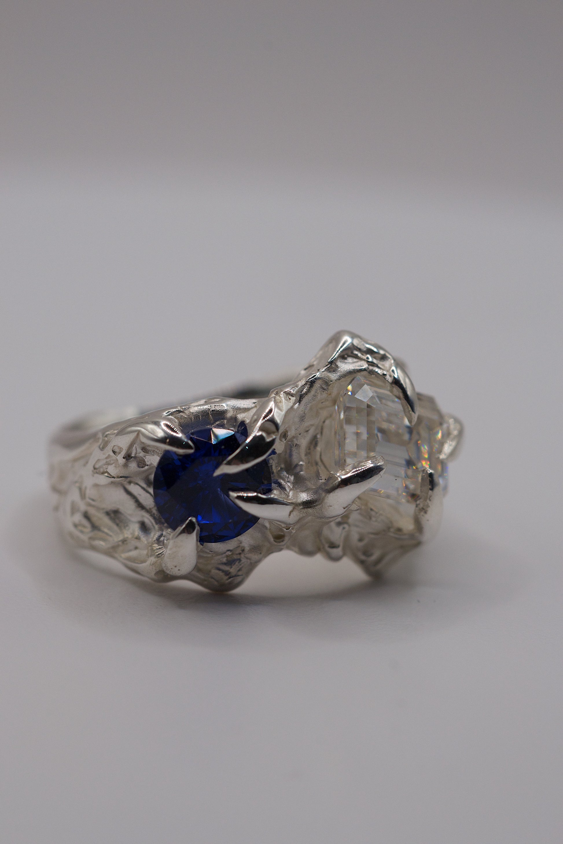 Handmade sterling silver alternative lost wax ring with round blue sapphire and emerald cut moissanite held in place by sharp gothic claws.